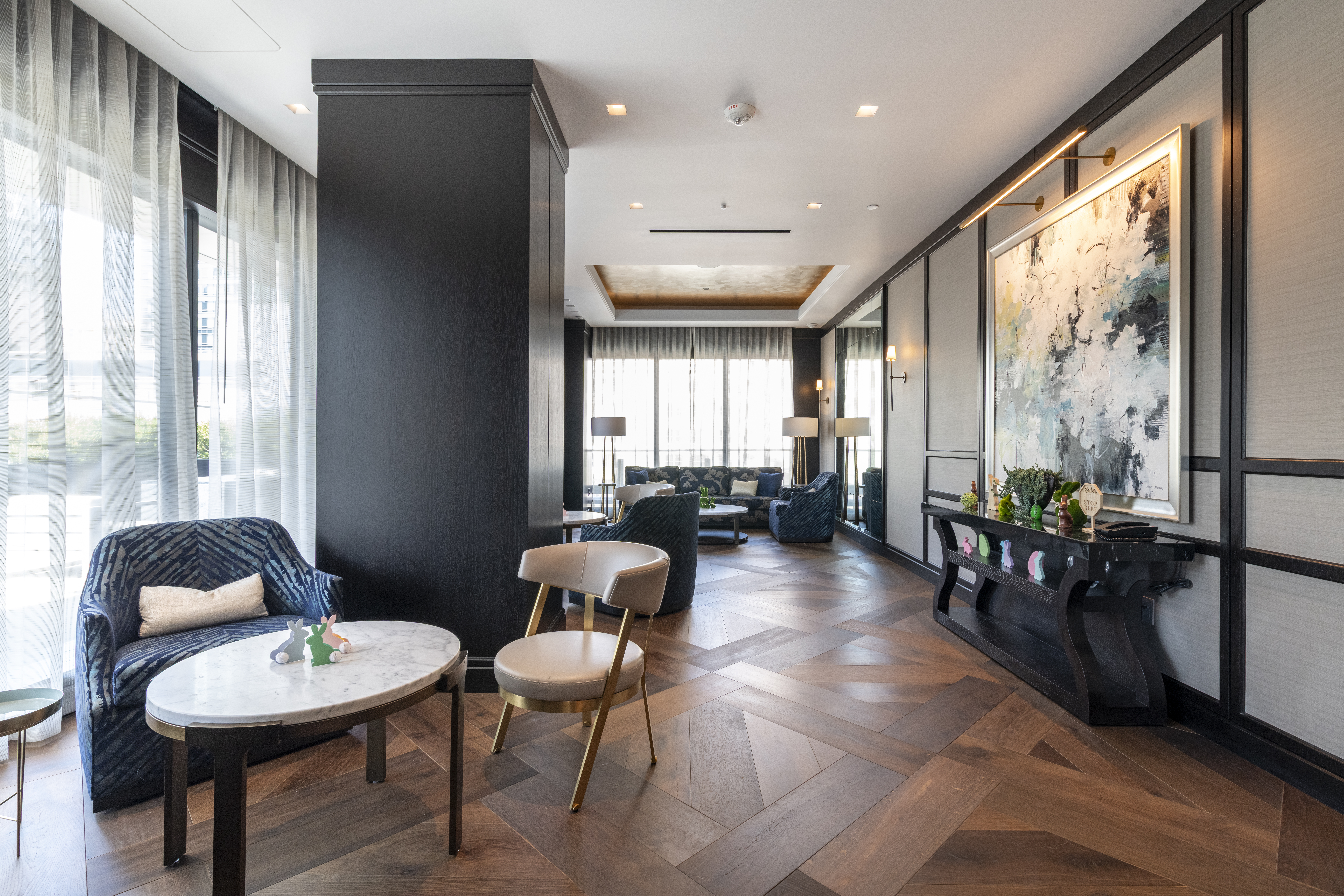 This sophisticated lounge area serves as an elegant community amenity, featuring a contemporary design with rich textures and a neutral color palette. High-end wood flooring, designer furniture, and curated art pieces create an upscale atmosphere, while large windows provide ample natural light. The space is thoughtfully arranged into multiple seating zones, making it an inviting area for social gatherings and relaxation.