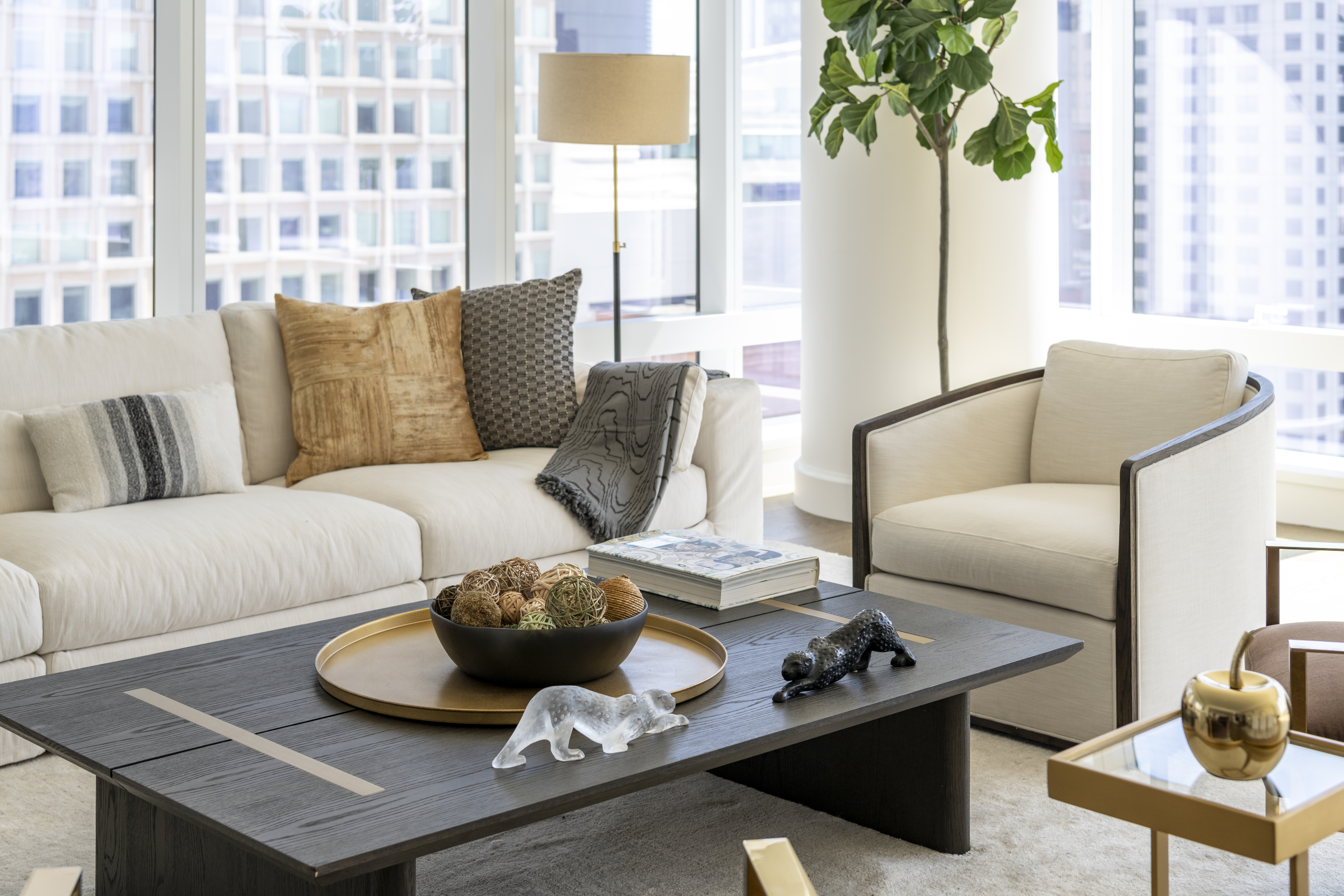 This sophisticated interior living space features upscale modern furniture, including a cream-colored sectional sofa accessorized with textured pillows and a charcoal throw. A dark wood coffee table serves as a focal point, styled with decorative spheres, a gold tray, and sculptural animal accents, all set against floor-to-ceiling windows that offer expansive cityscape views. A bright, airy atmosphere is complemented by a tall fiddle-leaf fig plant and elegant lighting, creating a luxurious and polished aesthetic.