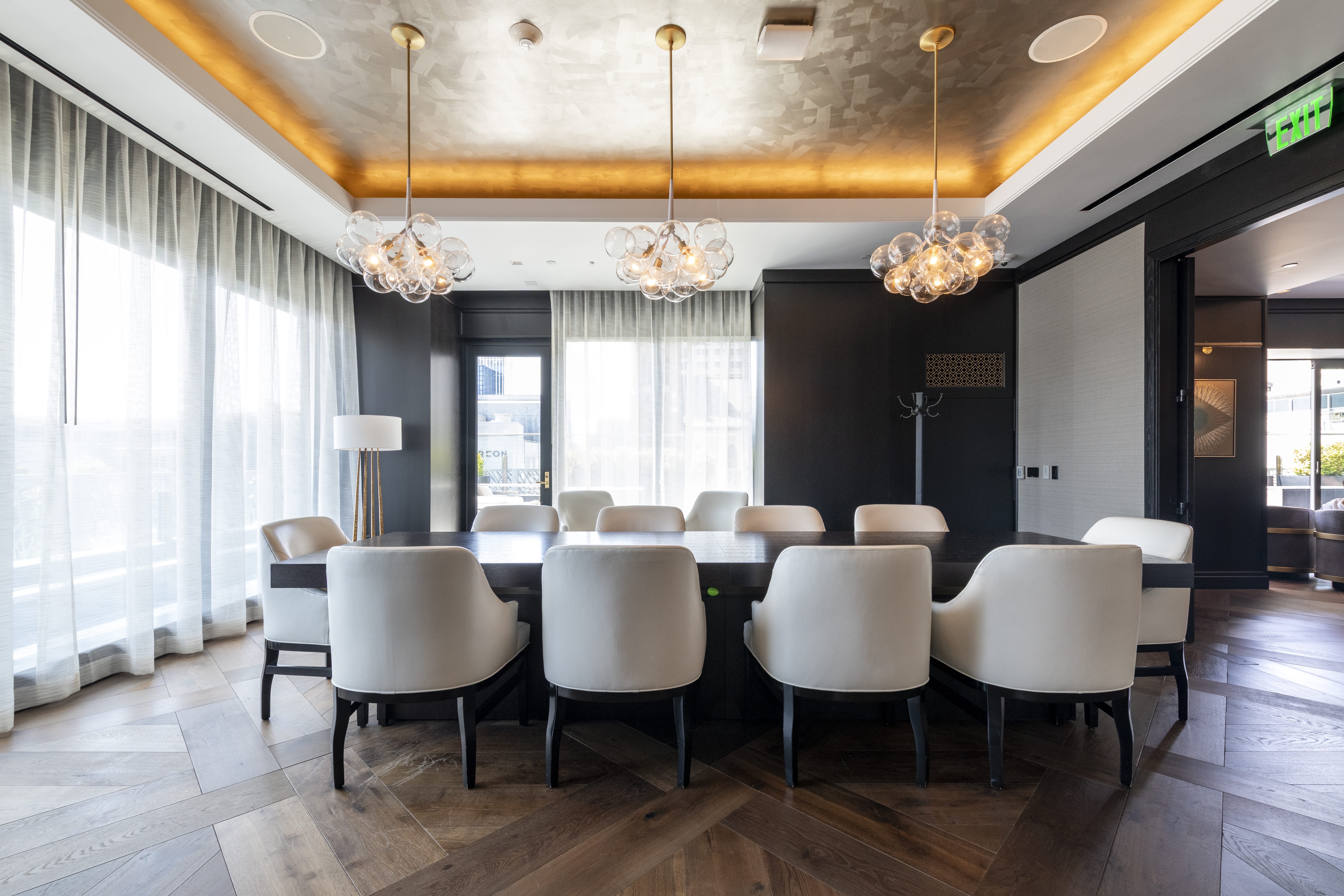 This sophisticated dining room features a long, dark wood table surrounded by elegant, upholstered white chairs. The space is illuminated by three distinctive glass globe chandeliers, set against a luxurious gold-leaf coffered ceiling and dark paneled walls. Large windows draped in light, sheer curtains allow for ample natural light, creating a bright and high-end formal dining atmosphere.