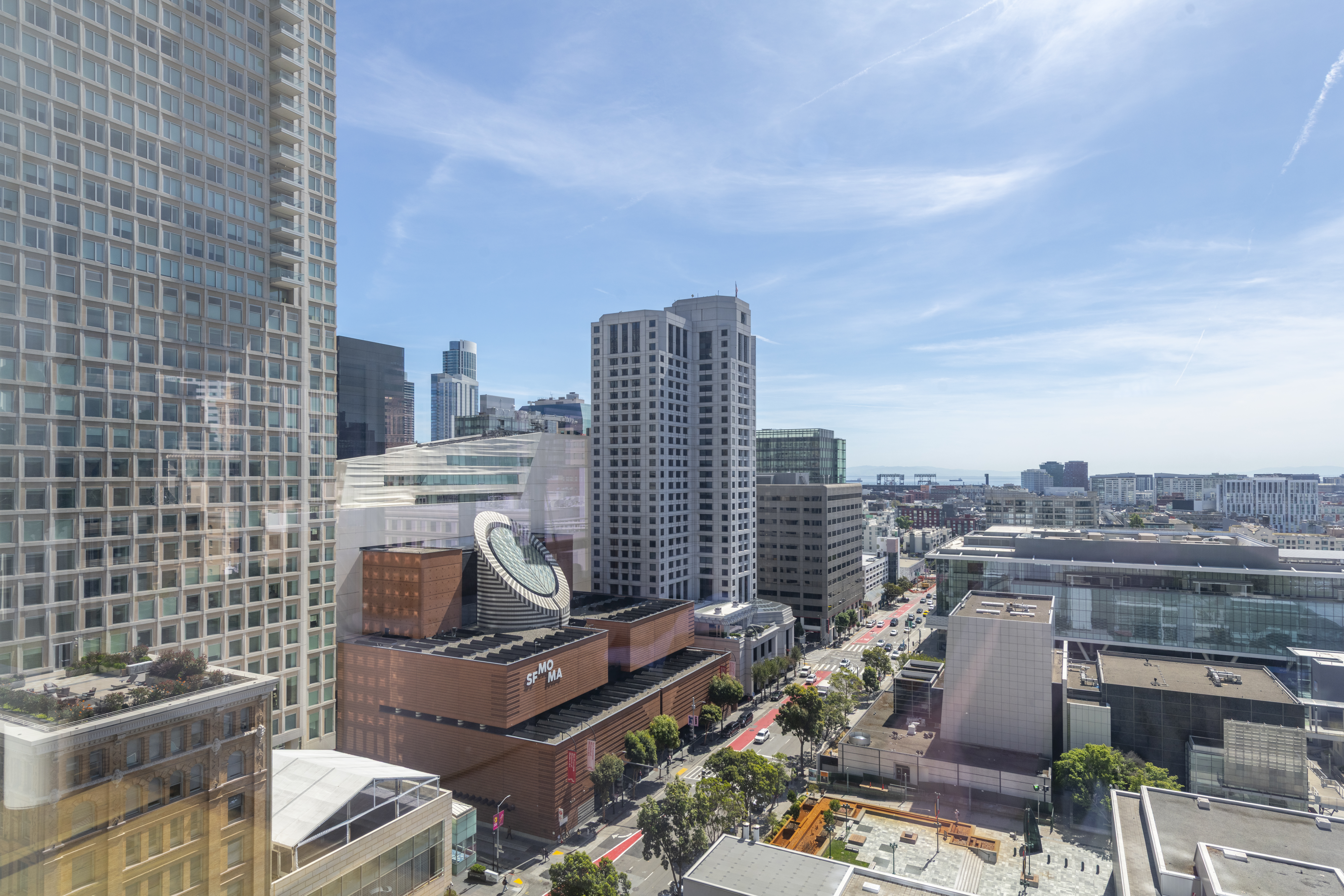 This high-angle, aerial perspective overlooks a dense urban cityscape, prominently featuring the SFMOMA museum building with its distinct brick facade and circular skylight. The scene captures a mix of modern skyscrapers, commercial office buildings, and street-level views of city traffic and greenery, offering a prime depiction of a metropolitan lifestyle. The bright blue sky and clear daytime lighting highlight the structural density and architectural character of this sought-after downtown location.