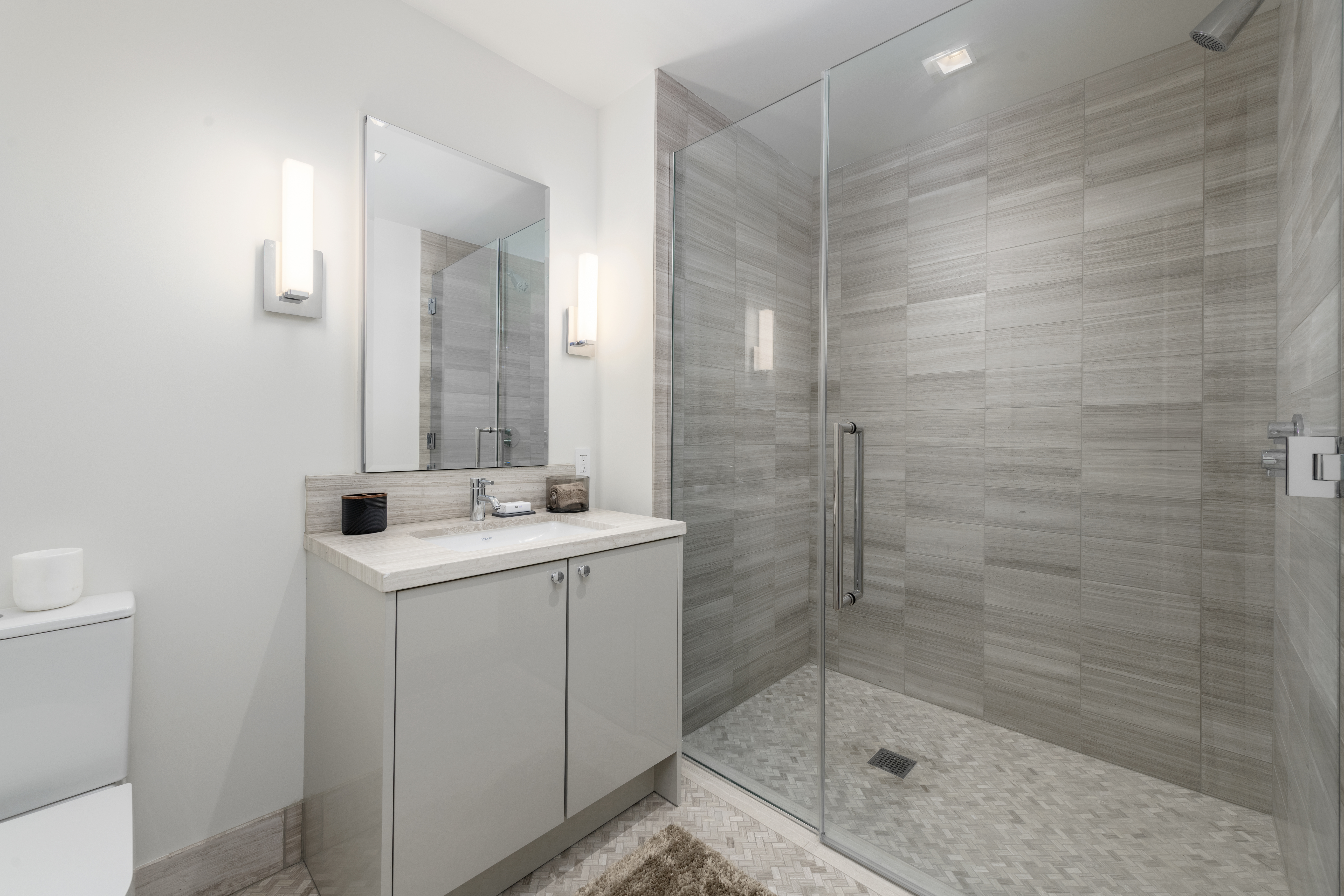 This modern bathroom features a sleek vanity with a stone countertop, a contemporary glass-enclosed shower with horizontal wall tiles, and refined lighting fixtures. The neutral color palette and clean lines create a polished, spa-like atmosphere suitable for a primary suite. The addition of patterned tile flooring in the shower enhances the room's upscale, sophisticated design.
