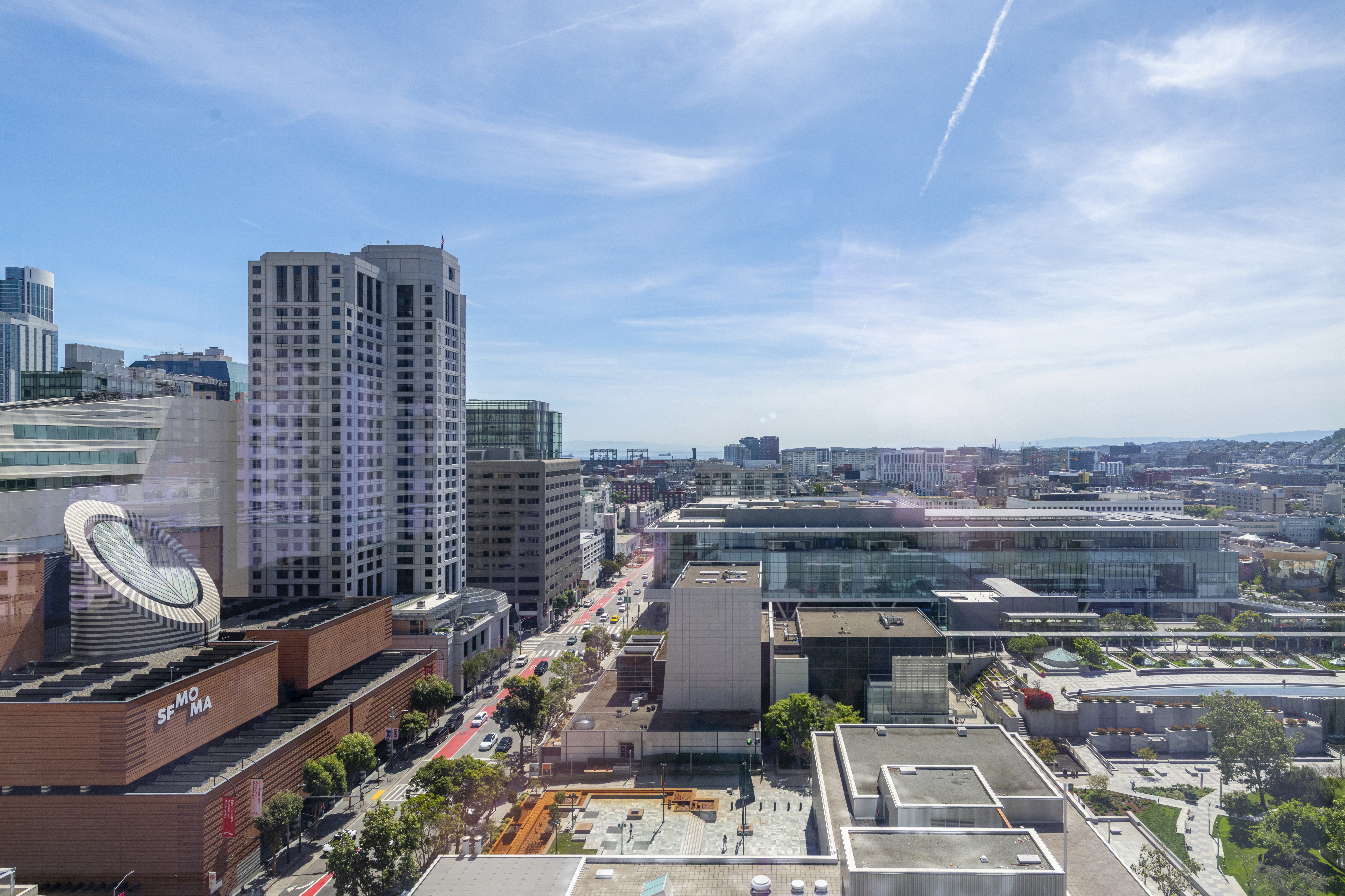 This high-rise aerial perspective captures a prominent metropolitan urban landscape, highlighted by the iconic SFMOMA museum building with its unique, modern architecture. The scene includes expansive city streets, large commercial office buildings, and public green spaces under a wide, clear blue sky. It provides a striking sense of the vibrant downtown neighborhood, perfect for showcasing a premier urban location.