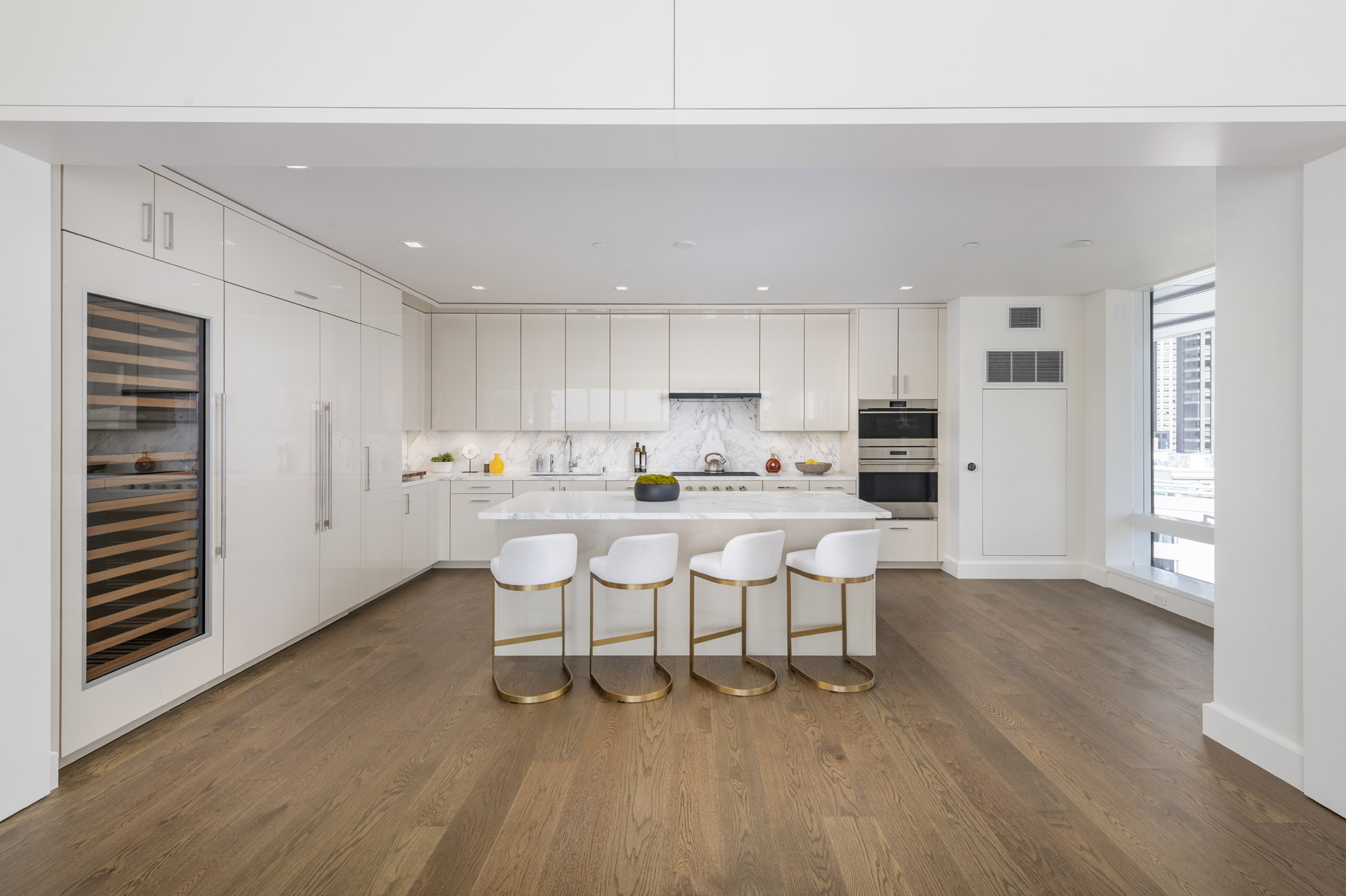 This sleek, contemporary kitchen features high-gloss white cabinetry, a large marble-topped island, and premium stainless steel appliances, including a built-in wine refrigerator. The open-concept design is enhanced by warm hardwood flooring and four stylish bar stools with gold metal bases, creating an inviting and luxurious space. Large windows provide bright natural light, emphasizing the immaculate, modern aesthetic throughout the room.
