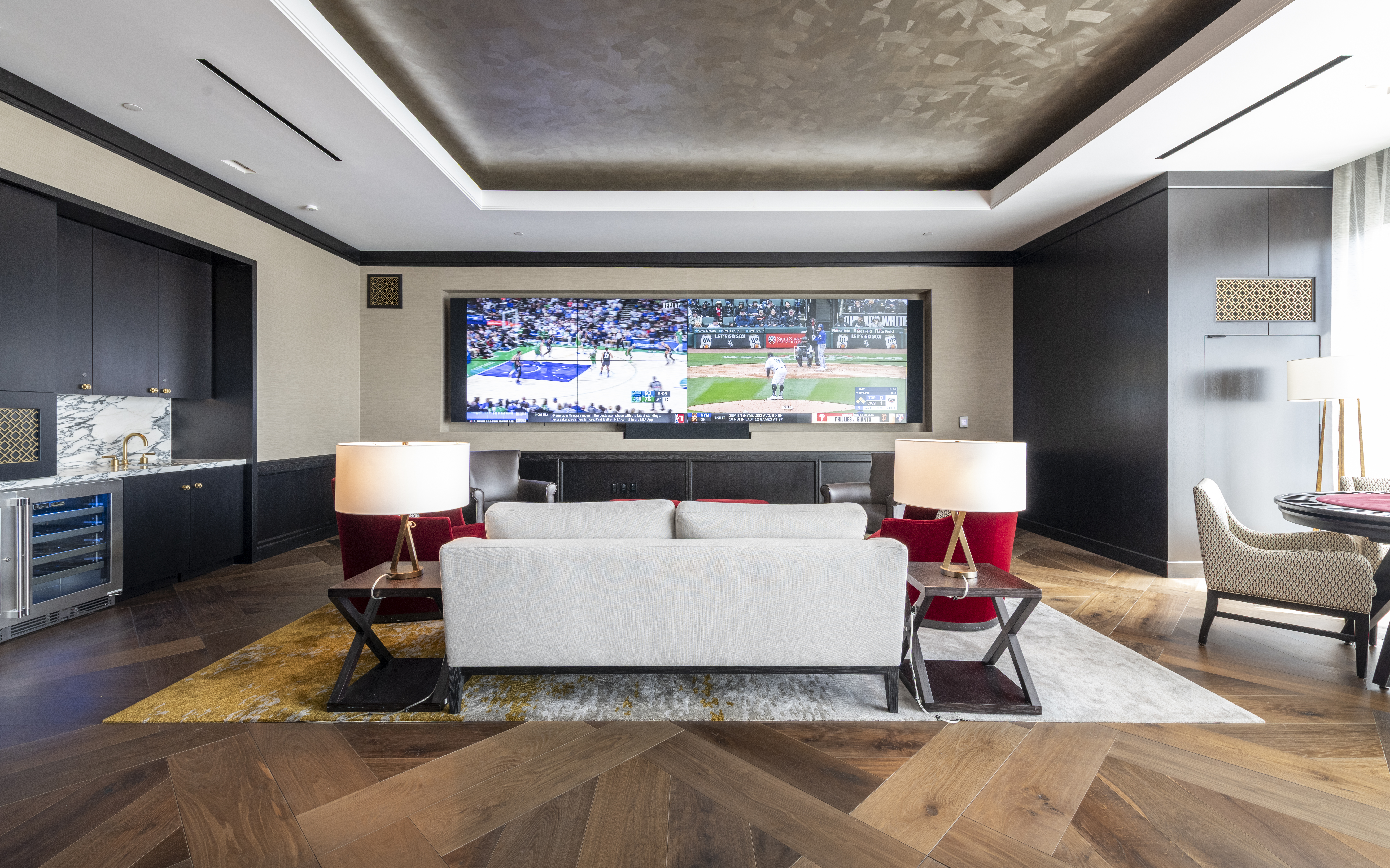 This sophisticated media lounge features a sleek, modern design characterized by a custom built-in entertainment wall showcasing a massive multi-screen display. The room is anchored by a contemporary cream-colored sofa, accented by red seating, and highlighted by rich herringbone hardwood floors and a statement metallic ceiling finish. A convenient wet bar with a wine cooler and marble countertop complements the space, creating an ideal setting for high-end entertainment.
