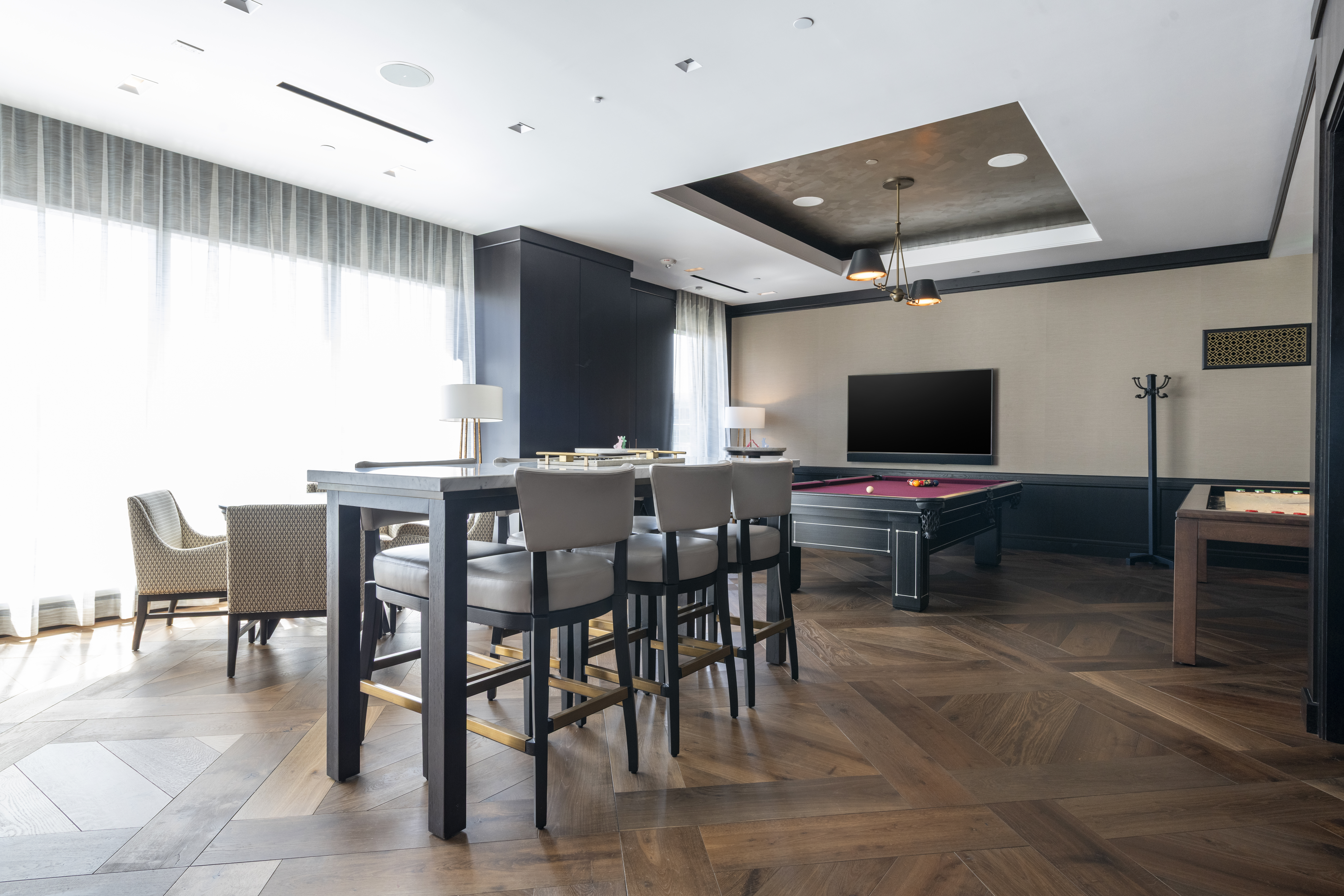 This sophisticated recreational room features a high-end billiards table, a bar-height table with stool seating, and an additional games table, making it an ideal gathering space for residents. The room is designed with rich wood chevron flooring, custom ceiling lighting, and elegant wall molding, creating an upscale and inviting atmosphere perfect for entertainment. Large floor-to-ceiling windows with soft drapery fill the space with natural light, emphasizing its luxurious and polished aesthetic.