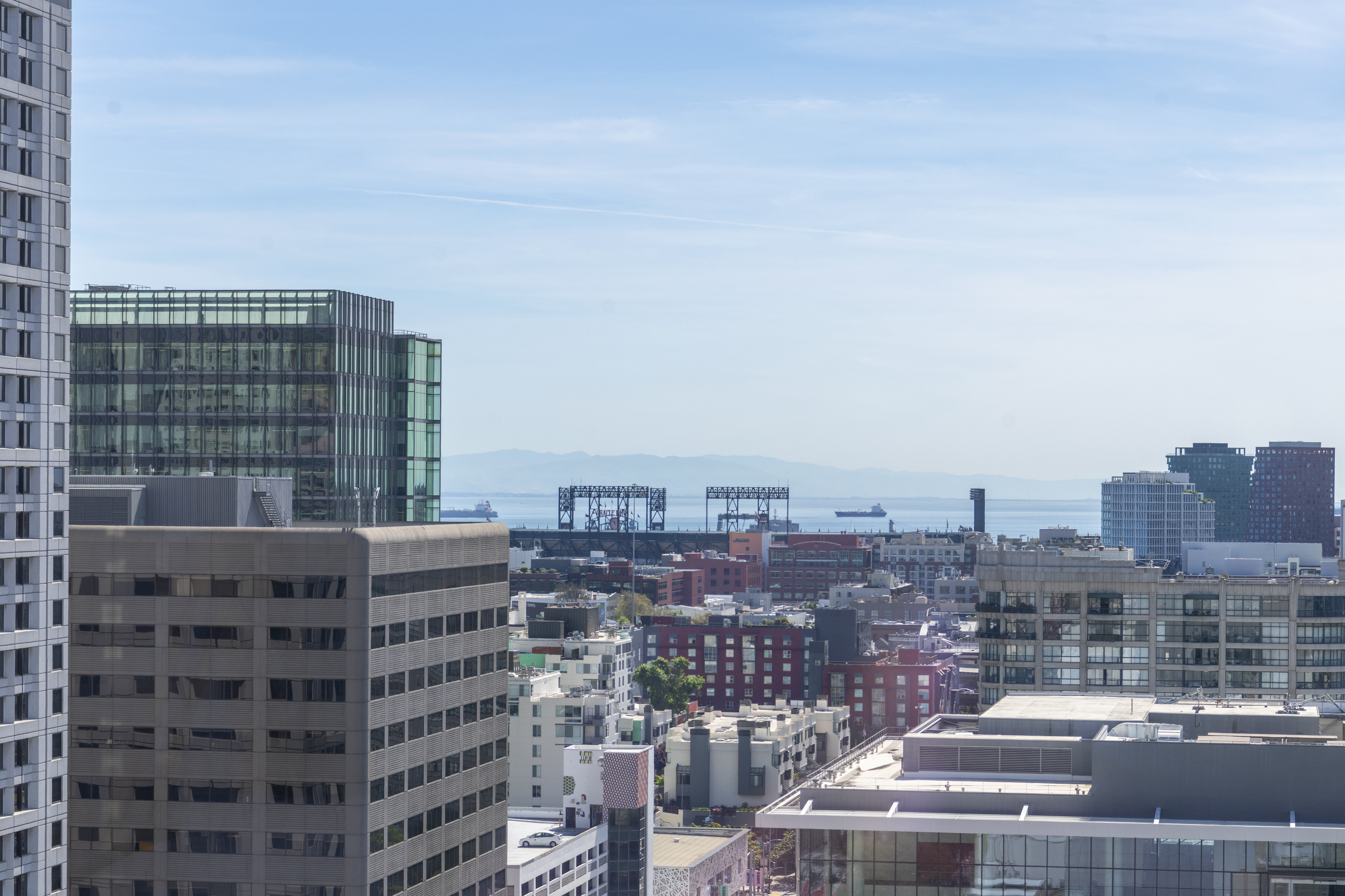 This high-altitude urban view captures a dense cityscape featuring a mix of modern glass high-rises and established mid-rise buildings. The perspective highlights the skyline's architectural variety, with the distant bay, container cranes, and mountains providing a scenic backdrop. The image emphasizes the central location's proximity to regional landmarks and waterfront activity.