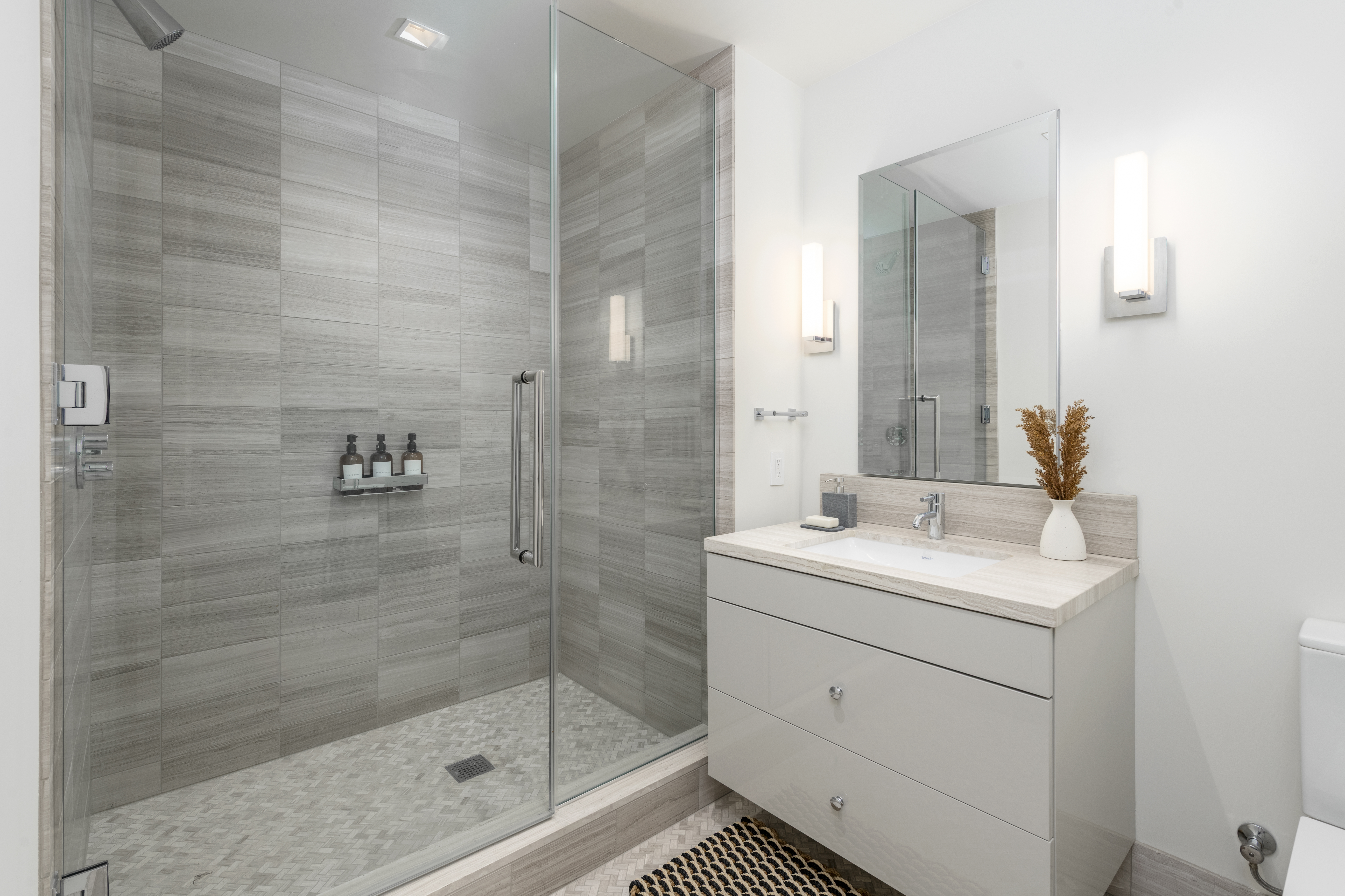 This modern primary bathroom features a spacious glass-enclosed walk-in shower with stylish horizontal gray tile work and a mosaic tile floor. A sleek vanity with a stone countertop and double drawers provides ample storage, complemented by sophisticated wall lighting and a large mirror. The overall aesthetic is contemporary, clean, and spa-like, making it an appealing feature for potential buyers.