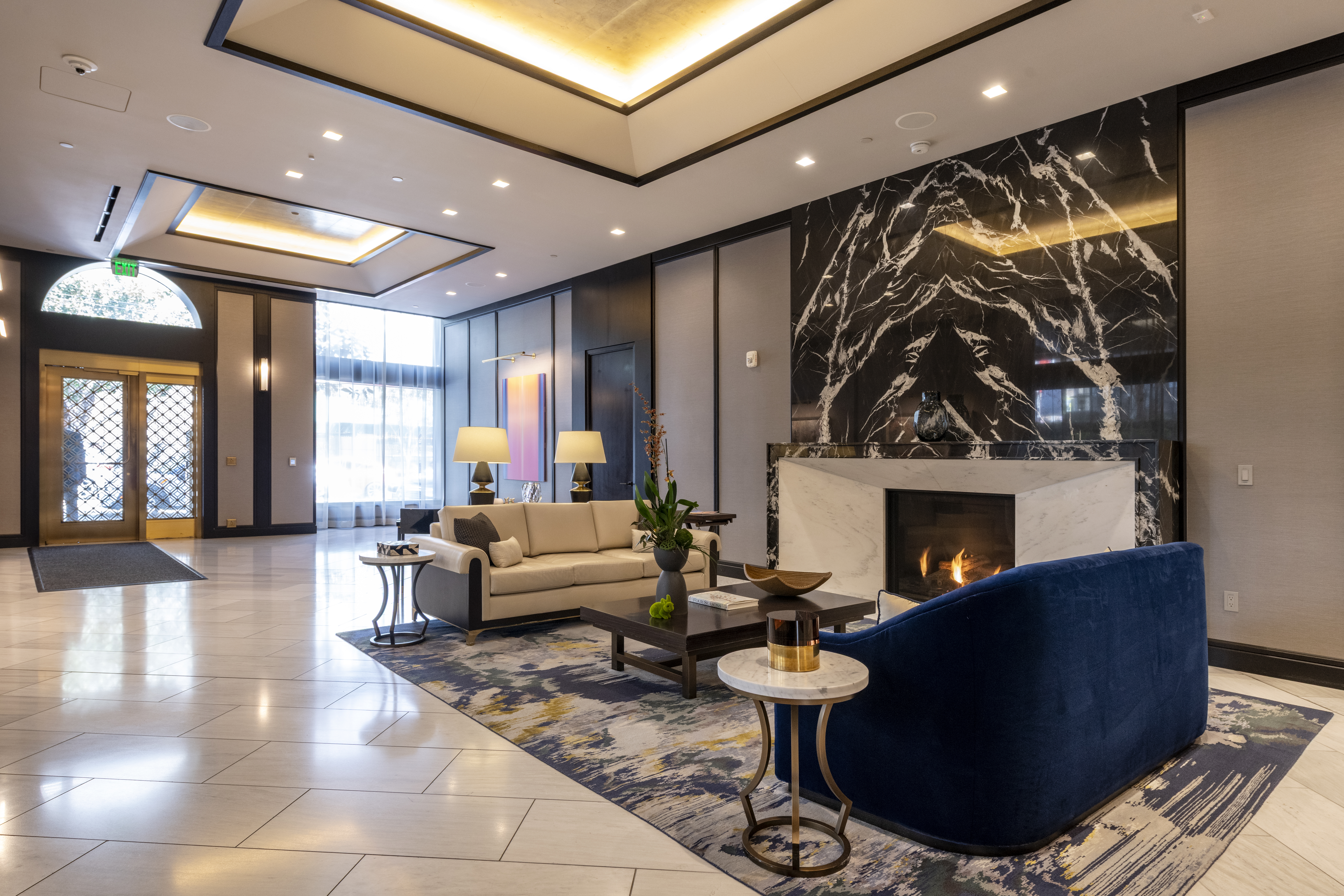 This sophisticated lobby or common area features a stunning marble-clad fireplace, plush navy blue and cream seating, and elegant designer lighting. The space exudes a luxury hotel atmosphere with its polished tile flooring, grand architectural ceiling details, and contemporary artwork. It is an inviting and upscale communal space designed to impress residents and guests alike.