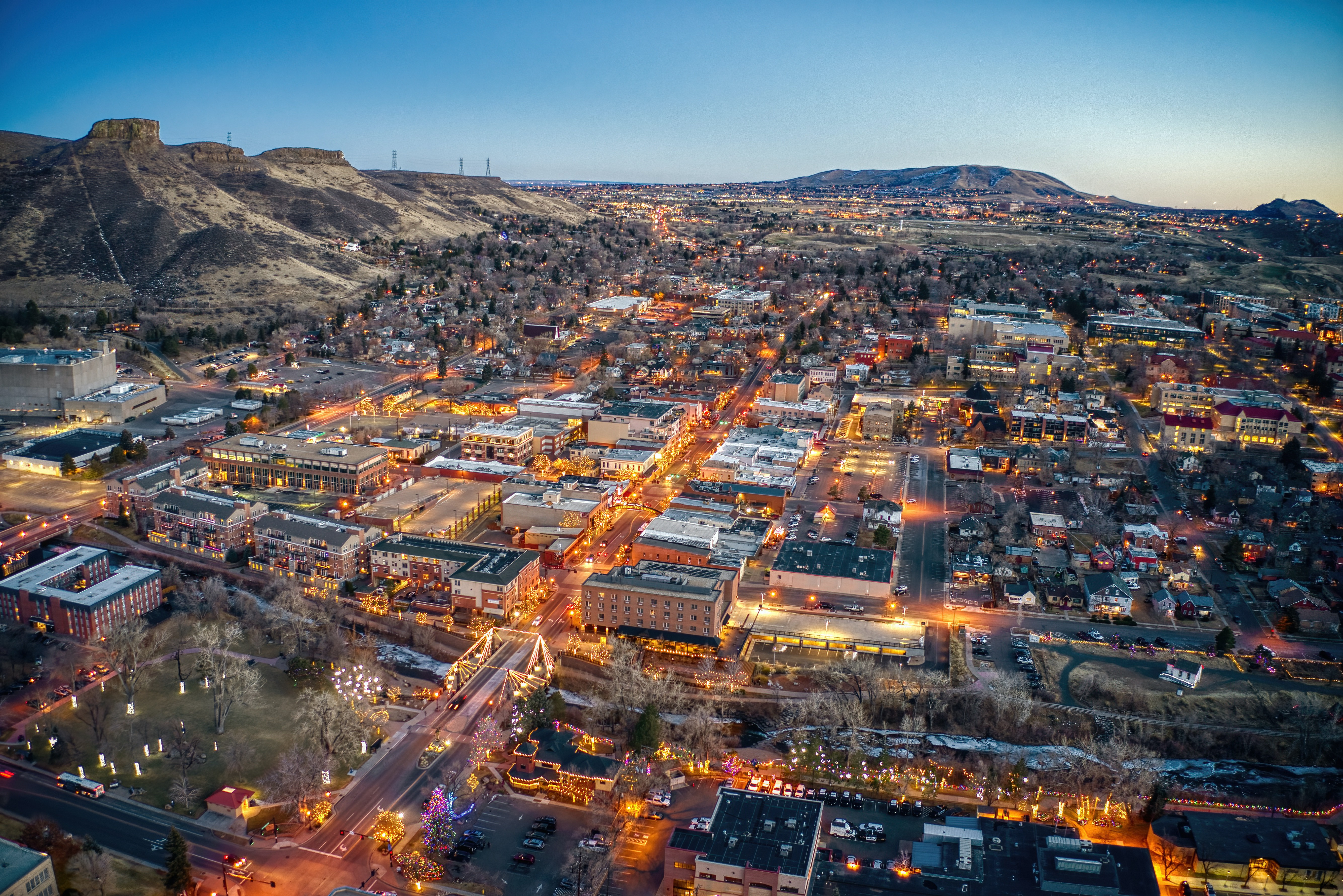 This aerial view showcases a vibrant town nestled against a scenic mountain backdrop at dusk. The well-lit streets and buildings suggest a bustling atmosphere, with a mix of commercial and residential properties. A prominent bridge adds character, and the overall impression is of a charming and well-maintained community.