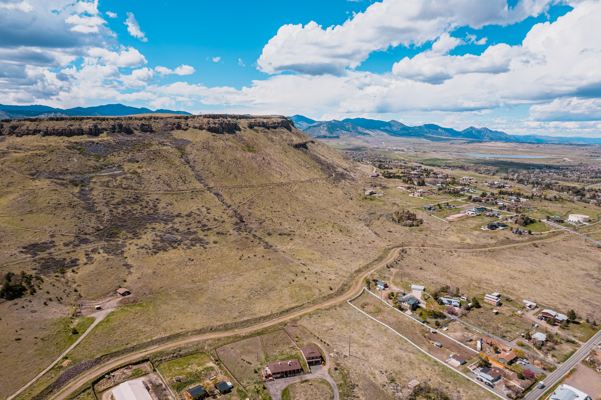 This aerial view showcases a vast landscape featuring a mesa prominently in the foreground. The scene transitions into a residential area with houses and streets, stretching to distant mountains under a bright blue sky dotted with fluffy white clouds. This shot would highlight the property's proximity to nature and community.