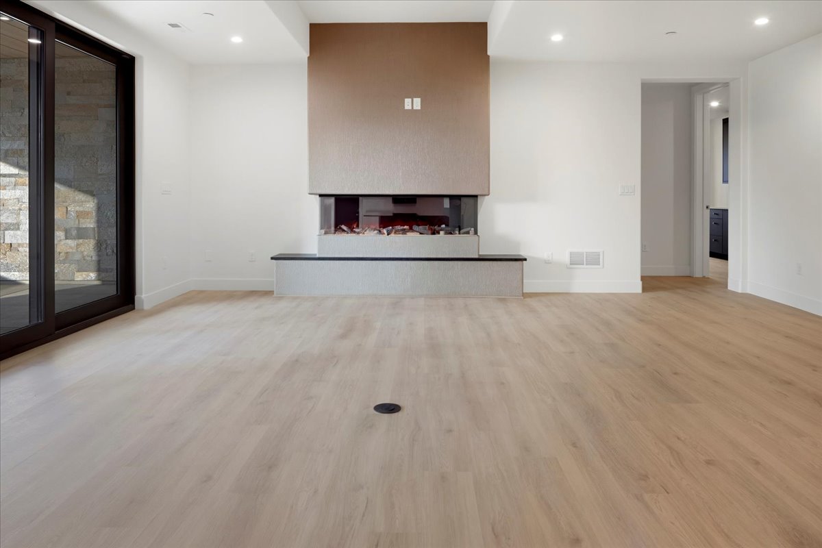This is a spacious living room featuring a modern fireplace with a unique design and a neutral color palette. The light-colored hardwood flooring and white walls create a bright and airy atmosphere. A large sliding glass door provides natural light and possibly access to an outdoor area, enhancing the room's appeal.