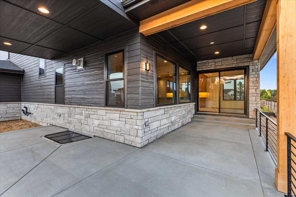 This image showcases a modern patio area featuring a gray concrete floor, stone accents, and dark gray siding. Large windows and doors provide access to the interior, while contemporary railings add a touch of style. The space appears to be designed for outdoor living and entertaining.