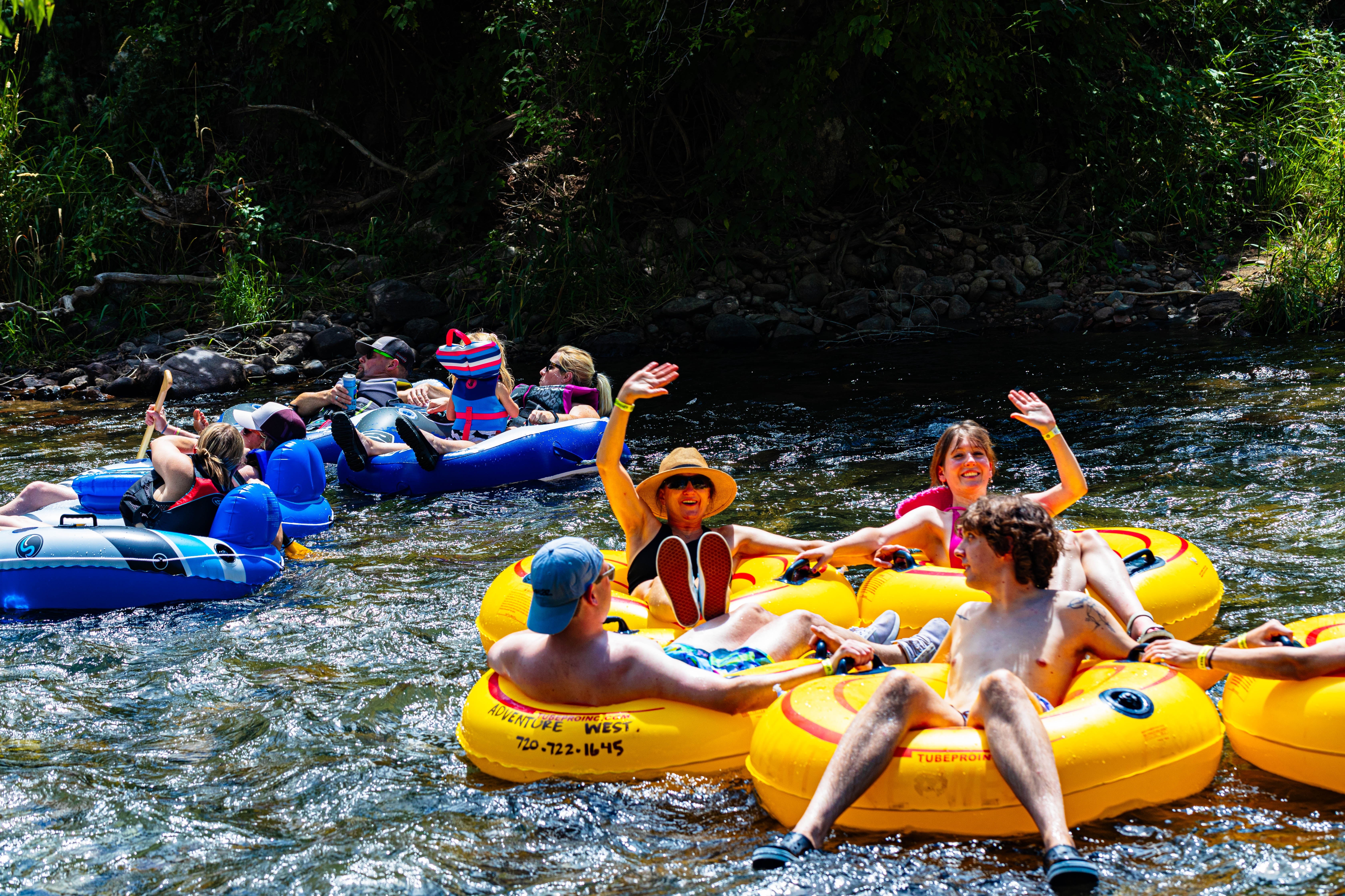 This image shows a group of people river tubing, an outdoor recreational activity. They are floating on inner tubes down a river, enjoying a sunny day. The scenery is natural, with trees lining the riverbank, possibly indicating proximity to a natural amenity.