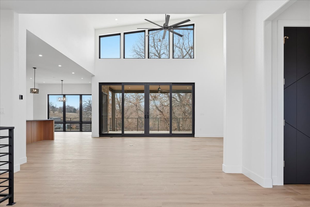 This is a bright and airy living room with high ceilings and expansive windows that offer natural light and views of the outdoors. The space features a modern design with minimalist decor, light wood flooring, and a neutral color palette, creating a spacious and inviting atmosphere. A ceiling fan and black framed windows and doors add to the contemporary look.