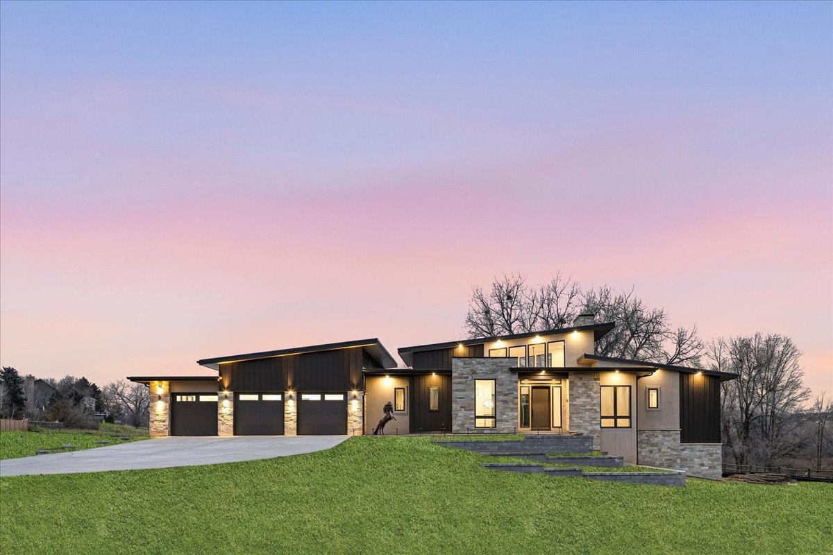 This is a front view of a modern home featuring a striking architectural design with a combination of stone and dark wood siding. The house presents a well-manicured lawn and a multi-car garage, creating a strong curb appeal. The property is illuminated by exterior lighting, highlighting the home's unique features, blending seamlessly with the colorful evening sky.