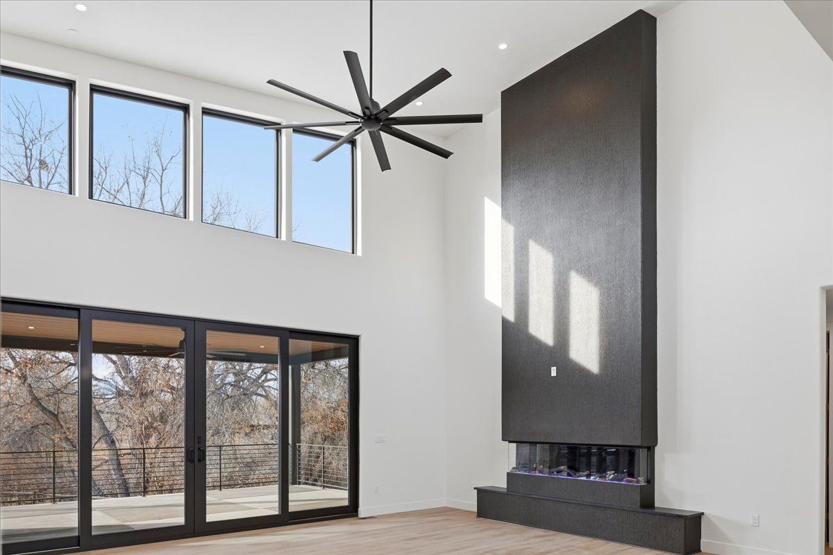 The image showcases a modern living room with an impressive two-story wall feature incorporating a fireplace. Large glass doors provide ample natural light and views of the outdoor space. A contemporary ceiling fan adds a stylish touch to the high ceiling, creating a grand and airy atmosphere.