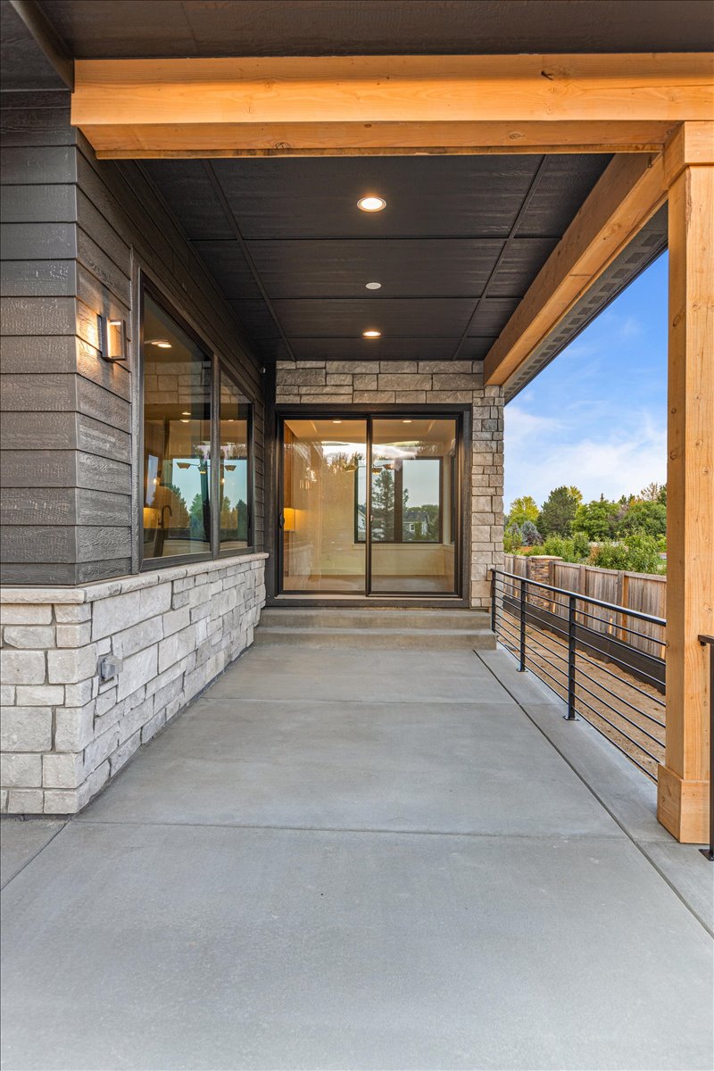 This image showcases a covered patio area with a concrete floor, stone detailing, and a modern railing. The space offers seamless indoor-outdoor living with a sliding glass door leading inside. Exposed wooden beams highlight the structure, and recessed lighting adds functionality.