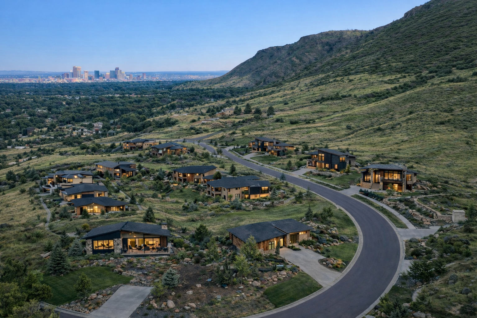 This aerial view showcases a luxury residential development nestled in a hillside setting. The modern homes feature dark roofs, expansive windows, and well-manicured landscaping that blends seamlessly with the natural surroundings. A winding road connects the homes, offering stunning views of the nearby city skyline.