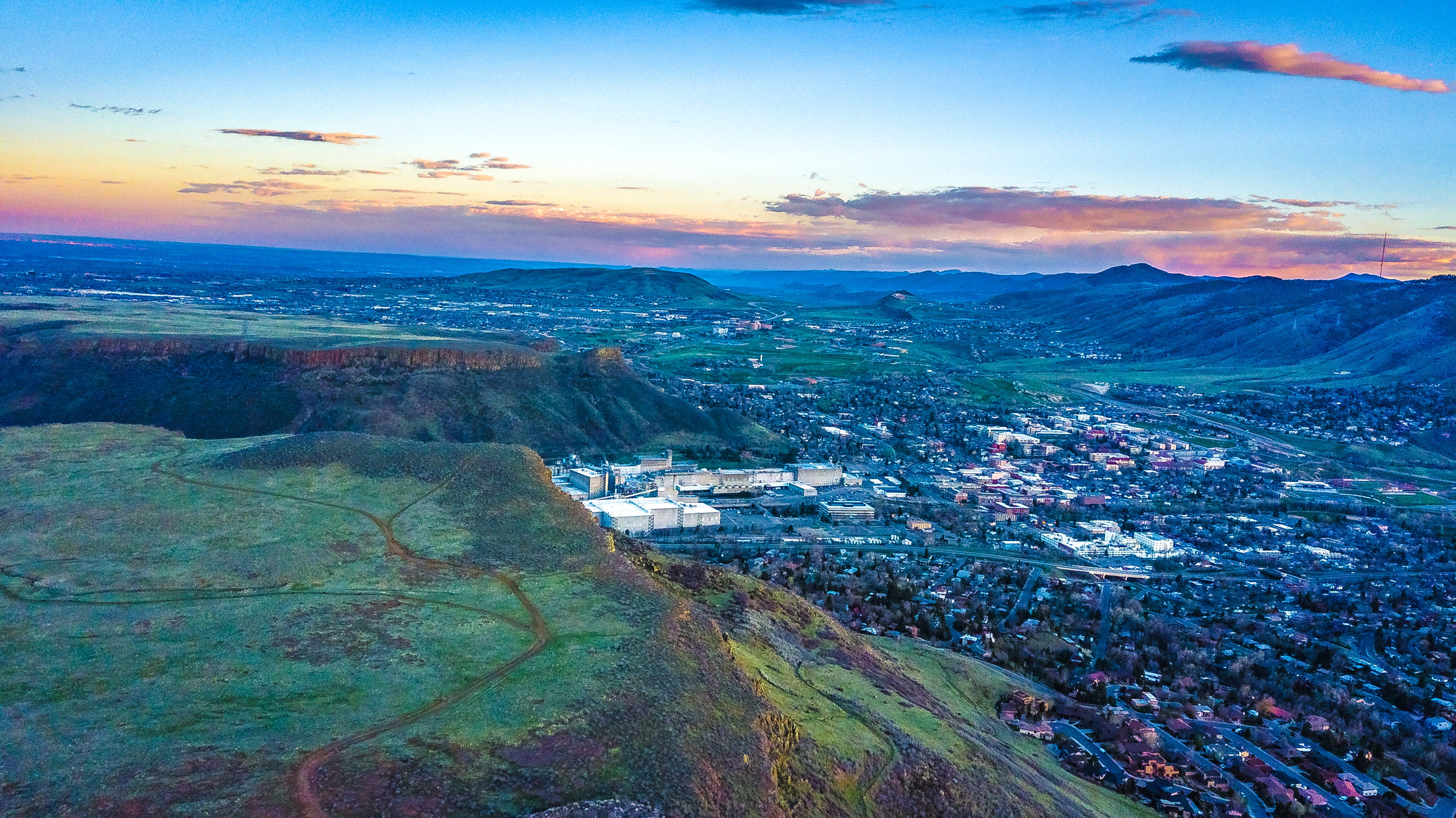 This aerial view showcases a sprawling landscape with a cityscape nestled between rolling hills and mountains. The foreground features lush green hillsides, giving way to a dense urban area, providing a sense of connectivity between nature and urban living. The sky is painted with hues of blue and soft pink, adding a tranquil element to the vibrant scene.