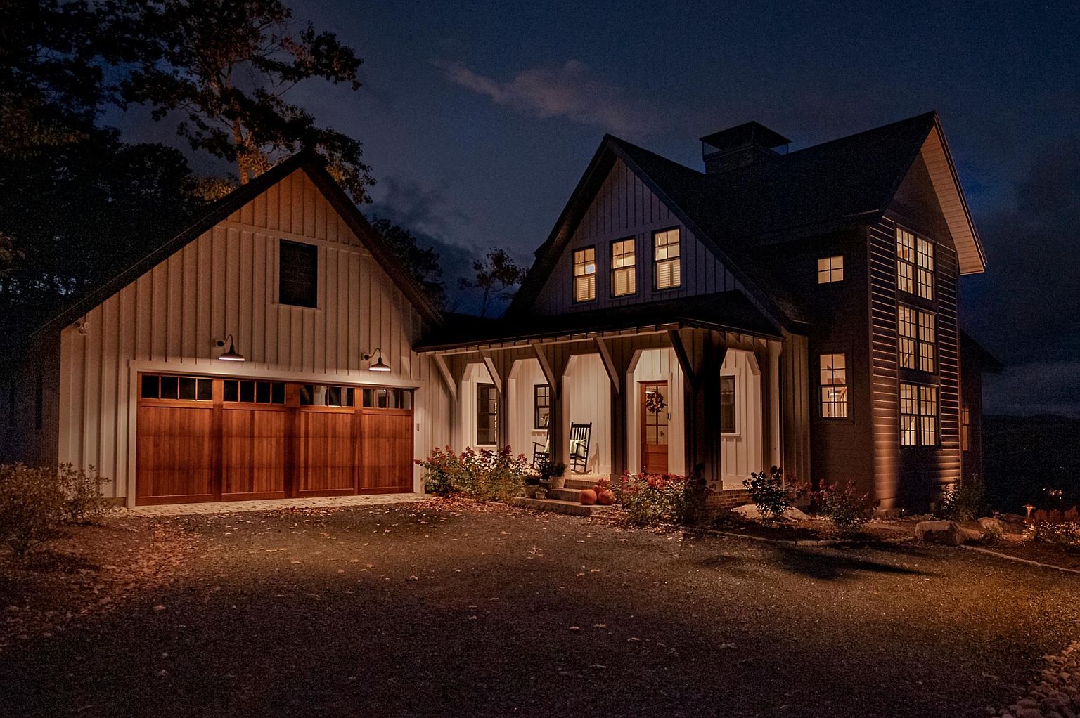 This charming farmhouse-style home is captured at dusk, showcasing a warm and inviting exterior with board-and-batten siding and a prominent attached garage. The front porch features decorative timber supports and a cozy seating area, while the warm glow from the interior windows creates a welcoming atmosphere against the twilight sky. The gravel driveway leads up to the structure, which is framed by mature trees and subtle landscaping, emphasizing a peaceful, high-end residential aesthetic.