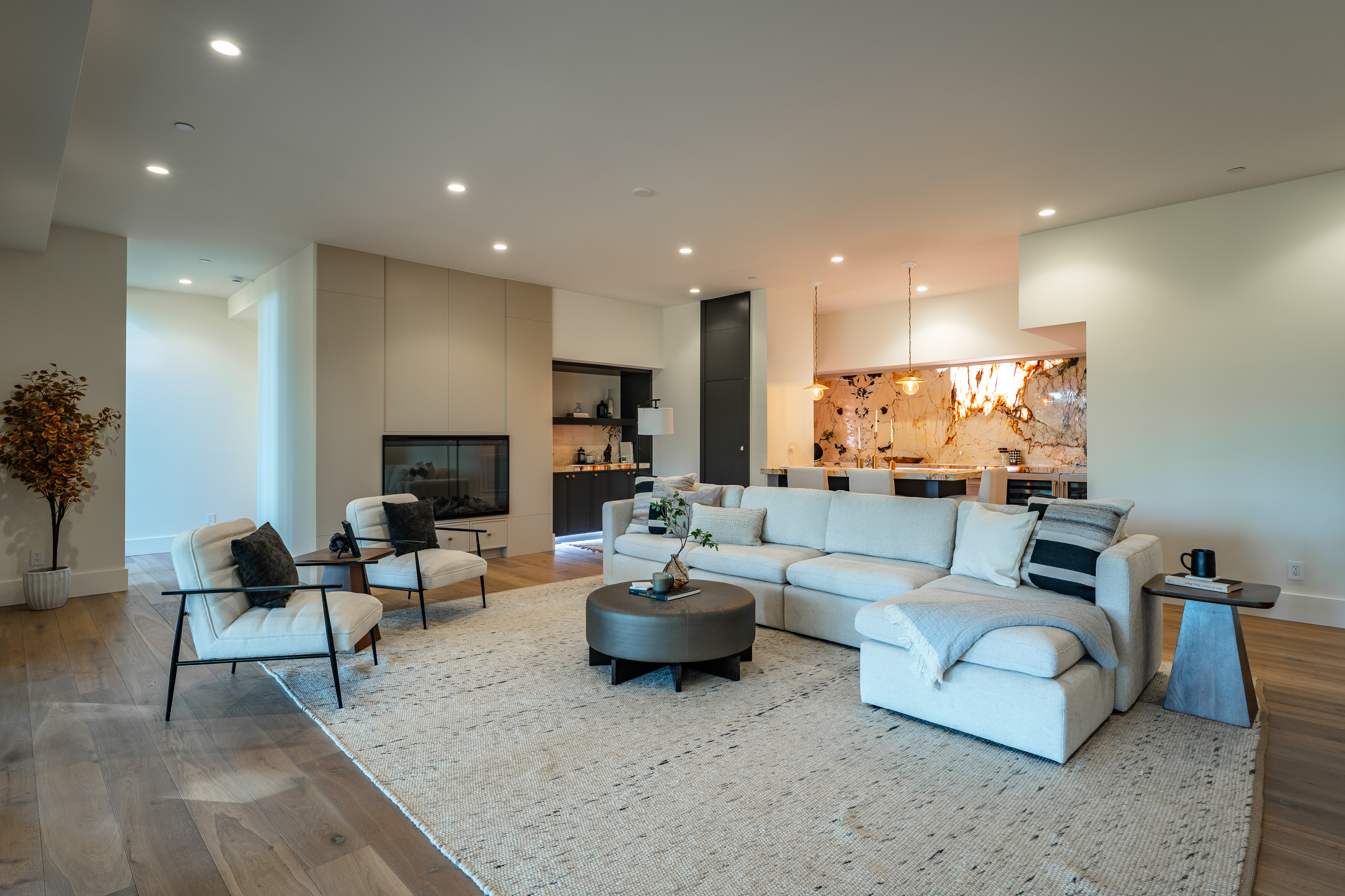 This is a bright and spacious living room featuring a large sectional sofa, comfortable armchairs, and a stylish area rug. The room boasts modern recessed lighting and a fireplace, providing a warm and inviting atmosphere. The open floor plan connects the living area to the kitchen, making it perfect for entertaining.