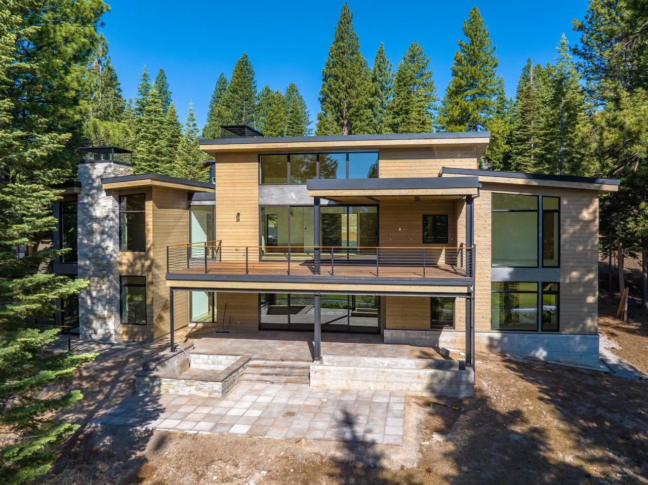 This image showcases the rear exterior of a modern two-story home, emphasizing its architectural design and outdoor living spaces. It features a large deck with metal railings and an expansive patio area below with a built-in fire pit. The house is nestled among tall trees, creating a private and scenic backdrop.