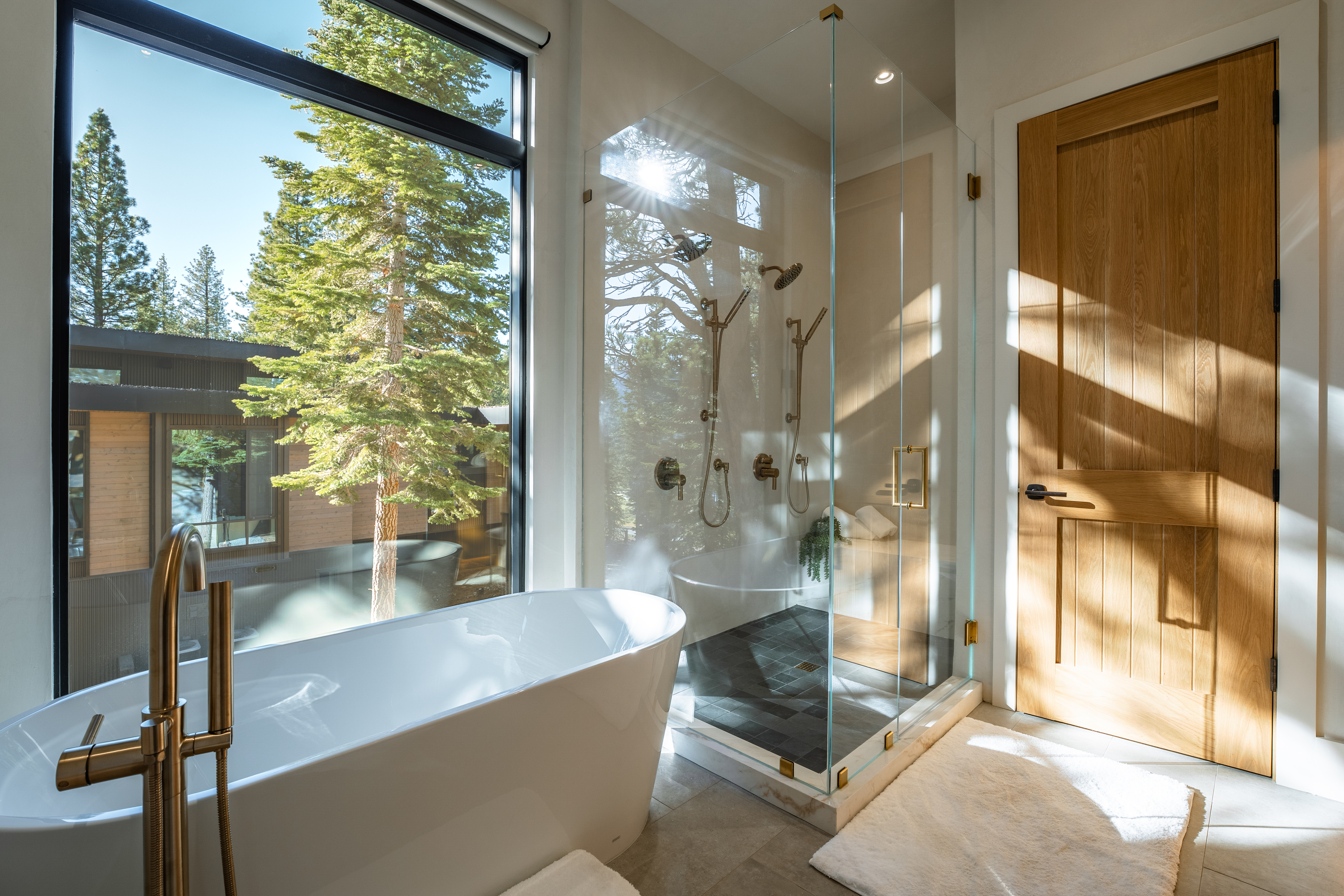 This is a beautifully designed primary bathroom featuring a freestanding tub with a golden faucet. Adjacent to the tub is a glass-enclosed shower with dual shower heads and black tiling, while a window provides a stunning outdoor view. A wooden door and plush rug complete the upscale and modern aesthetic of the space.
