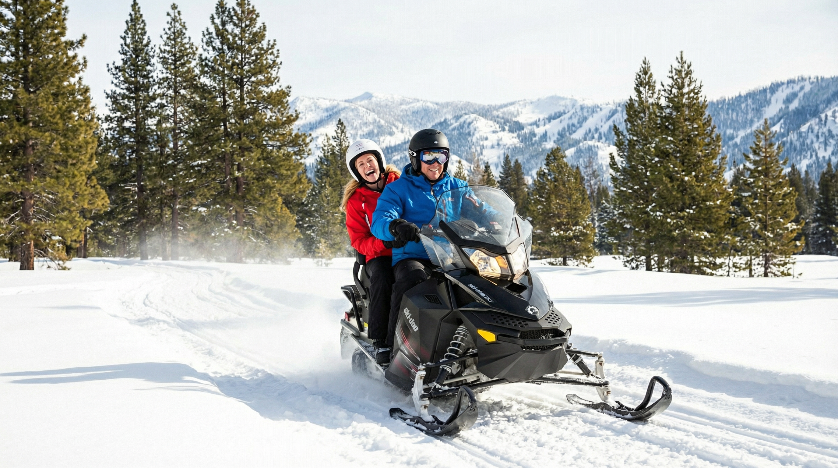This image showcases a couple riding a snowmobile through a snowy landscape with pine trees and mountains in the background. The scene highlights the recreational opportunities available in a winter setting. While not a traditional real estate photo, it could be used to market properties in areas known for winter sports and outdoor activities.
