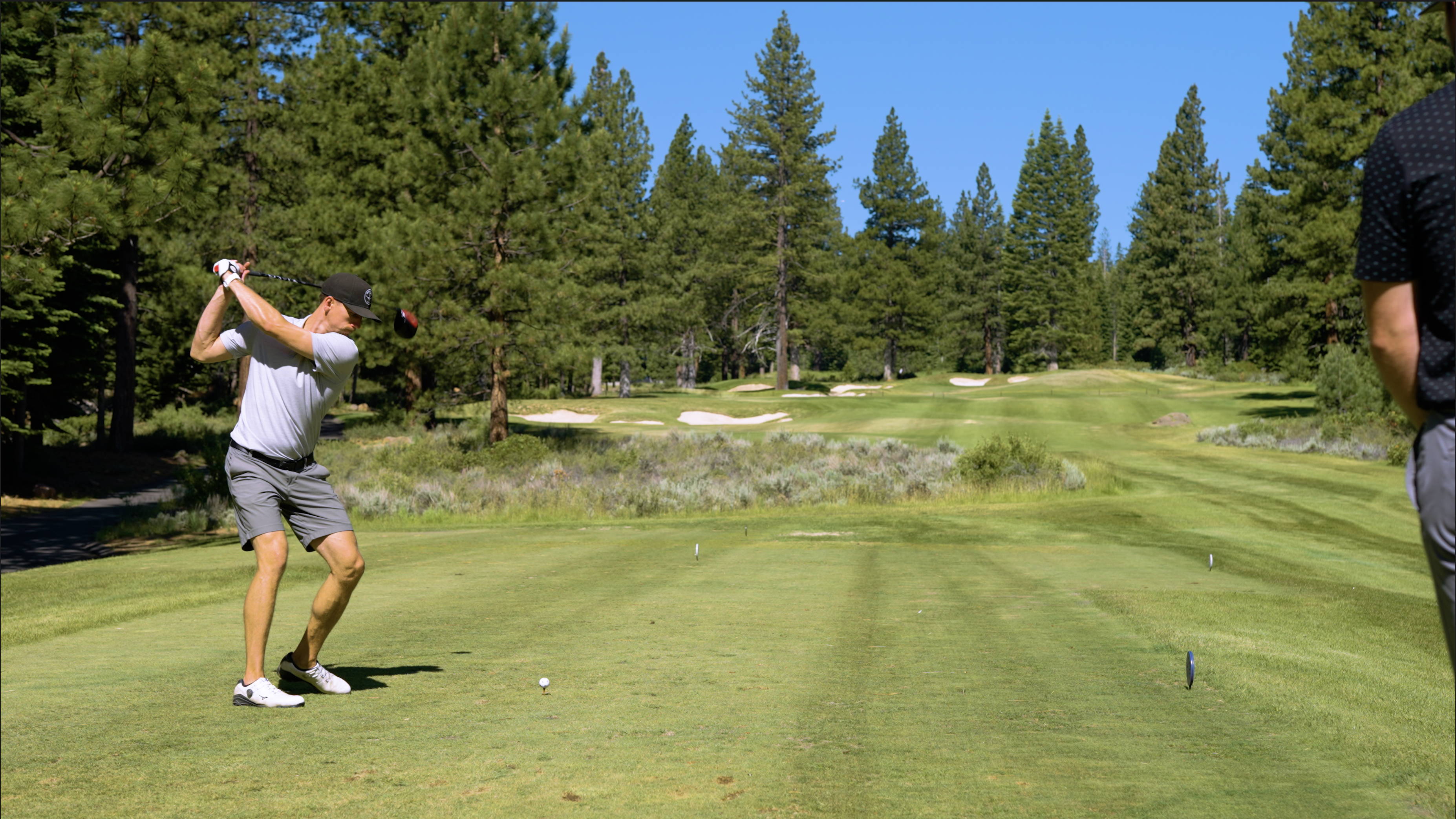 The image shows a well-maintained golf course with lush green grass, tall evergreen trees, and sand traps. A golfer is captured mid-swing, suggesting an active recreational environment. The scene promotes a sense of leisure and outdoor activities offered within the community or property.