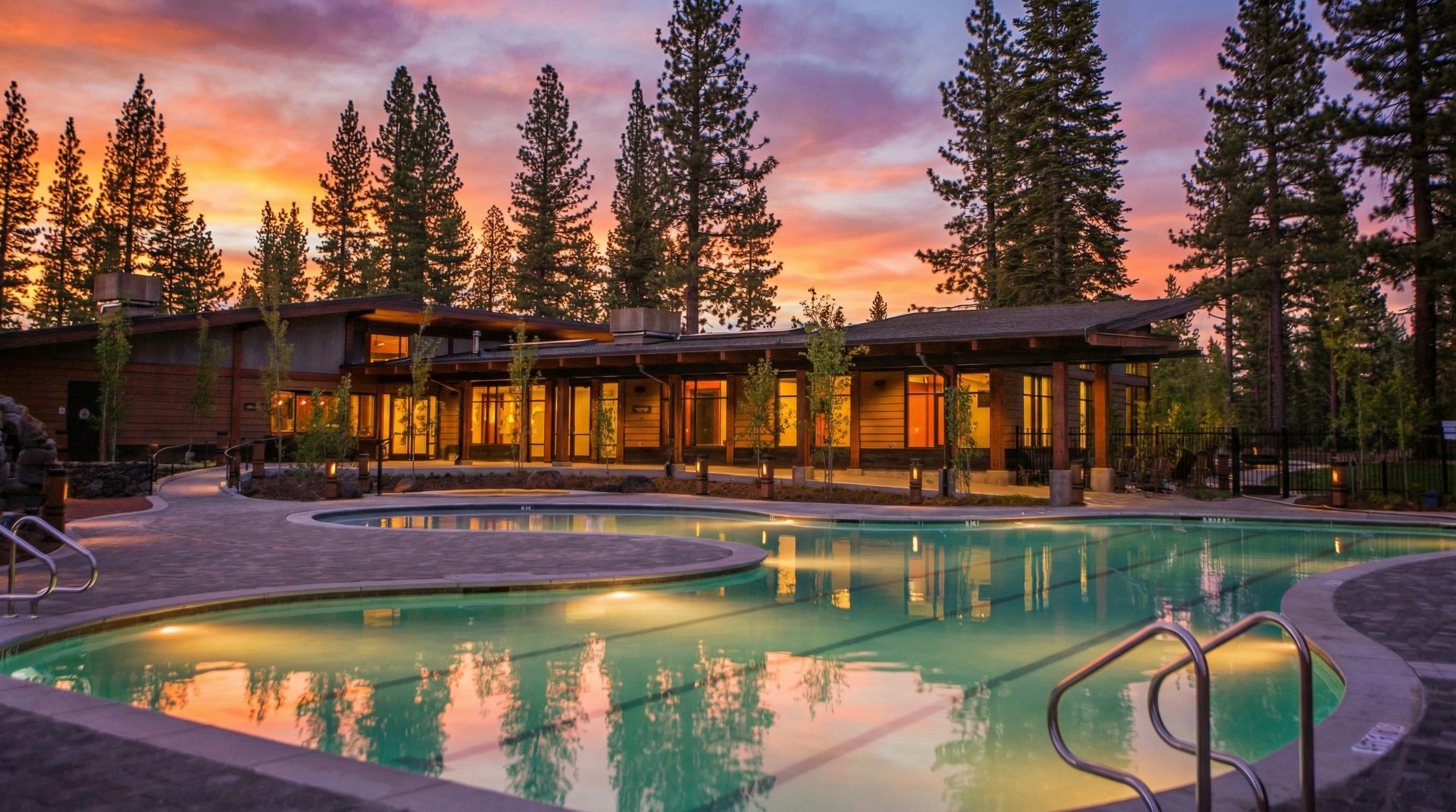 The image showcases a beautifully lit outdoor pool and spa area at dusk. The water reflects the warm glow of the lights and the colorful sunset sky. Tall trees surround the area, creating a serene and private ambiance, perfect for relaxation.