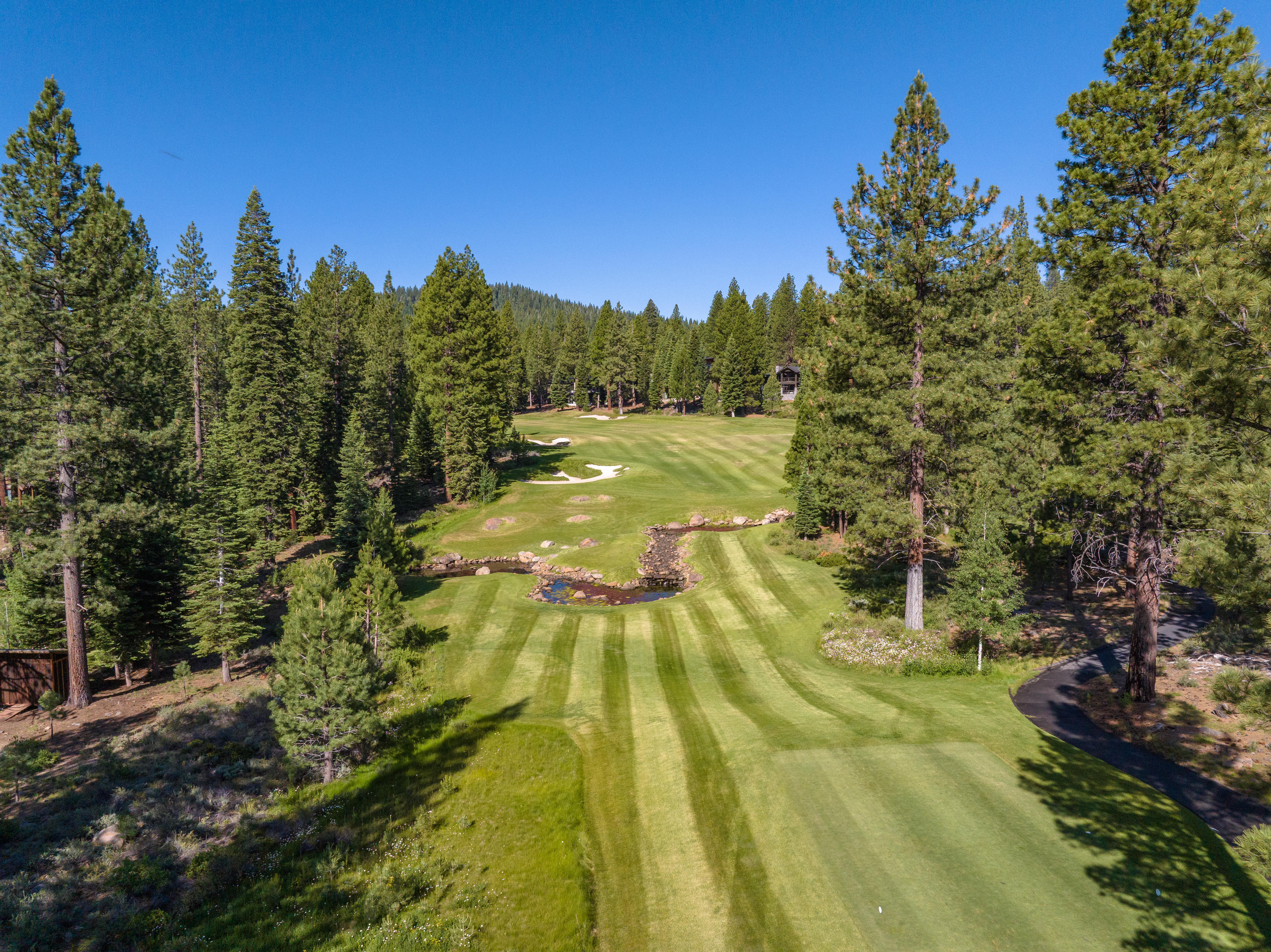 This aerial view showcases a pristine golf course bordered by lush evergreen trees under a clear blue sky. The manicured green fairways are punctuated by sand traps and a water feature, highlighting the course's design and maintenance. The surrounding trees add to the property's sense of seclusion and natural beauty, suggesting a serene and exclusive location.