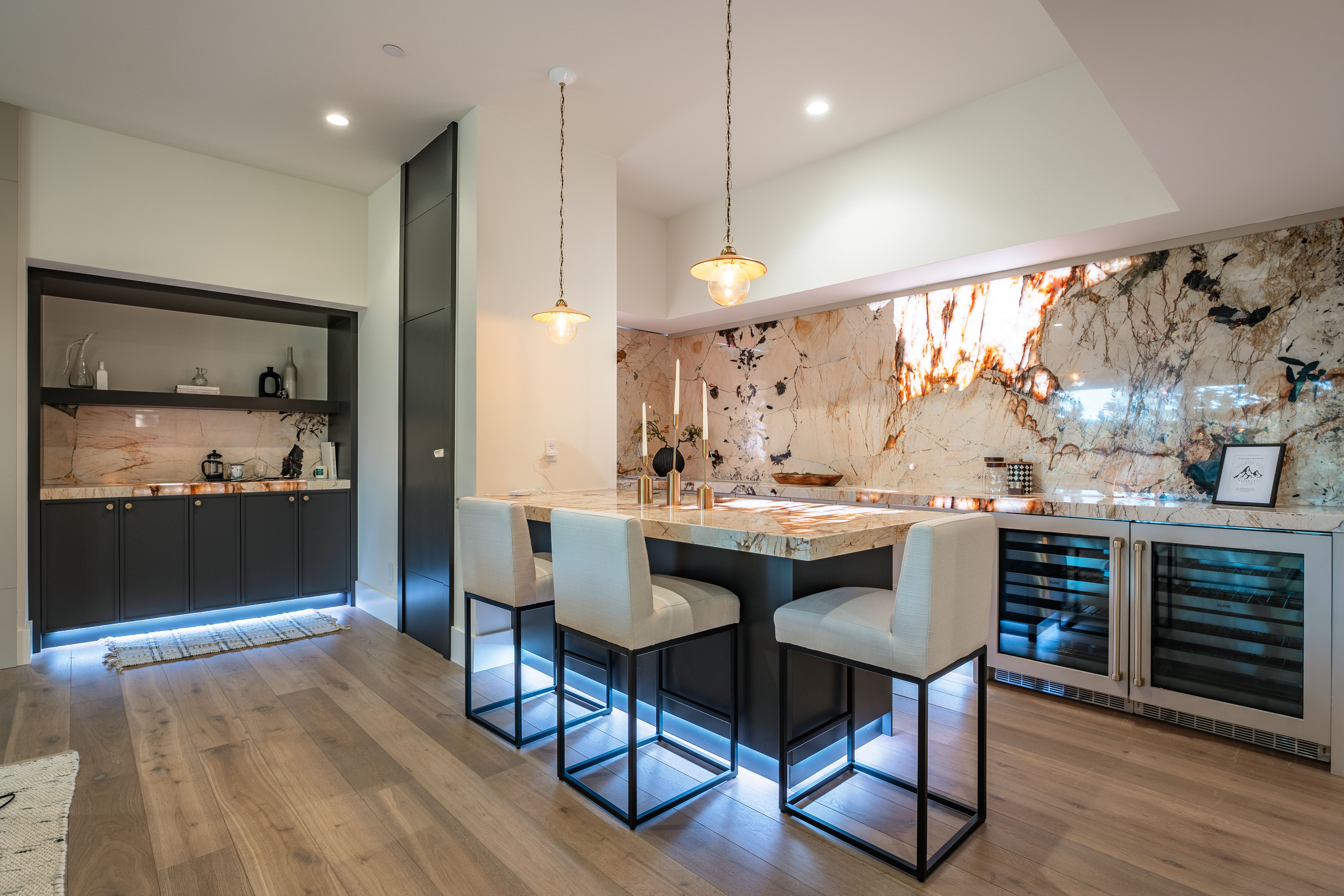 This image showcases a modern kitchen with a stunning illuminated marble backsplash. A large kitchen island with bar seating anchors the space, complemented by striking pendant lighting. Built-in wine storage adds a touch of luxury, contributing to a chic and sophisticated aesthetic.