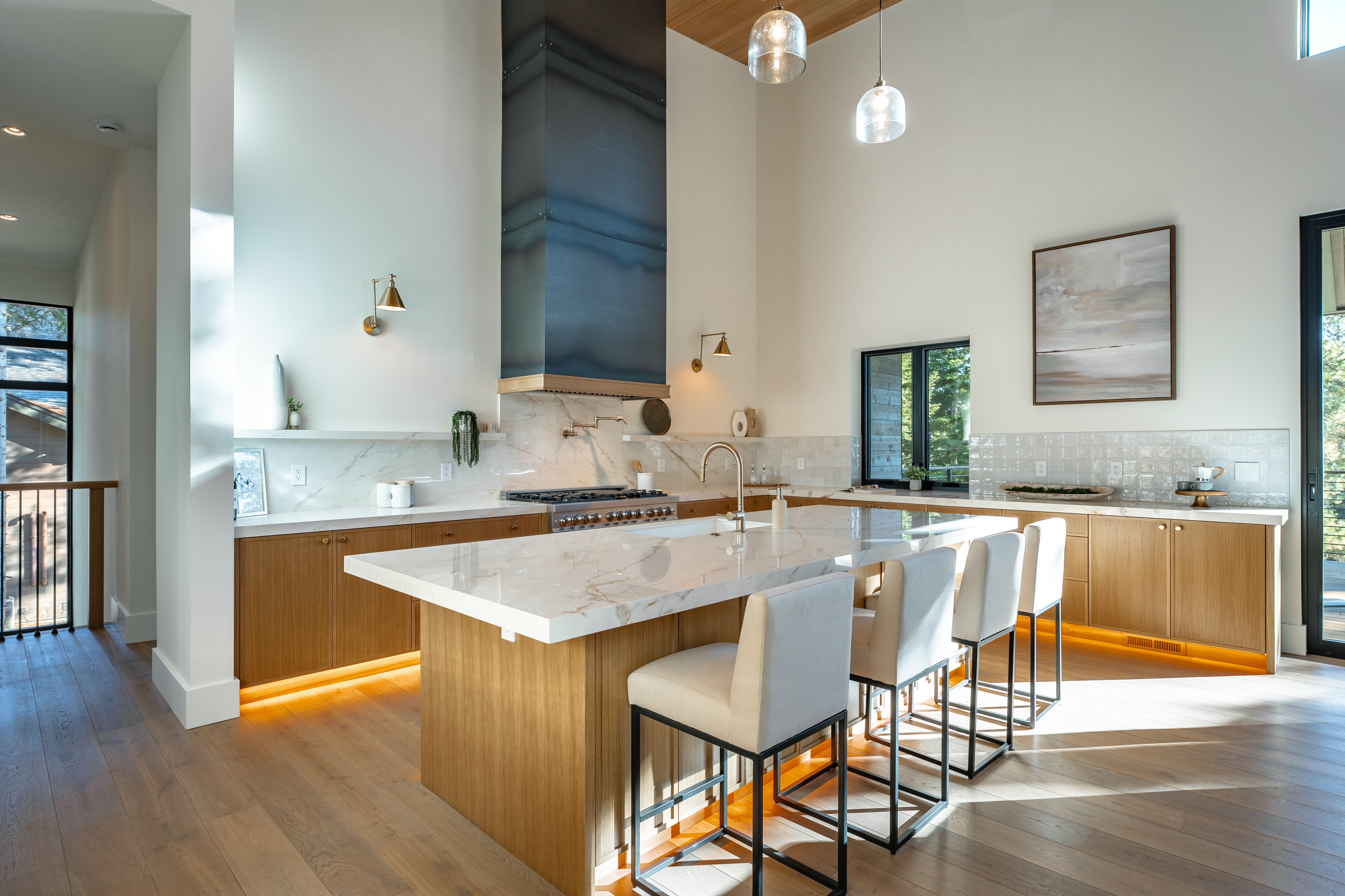 This is a bright, modern kitchen featuring wood cabinets, a large island with a marble countertop, and stainless steel appliances. Ample natural light floods the space, highlighting the hardwood floors and stylish bar seating. The kitchen is designed with an open layout and clean lines.