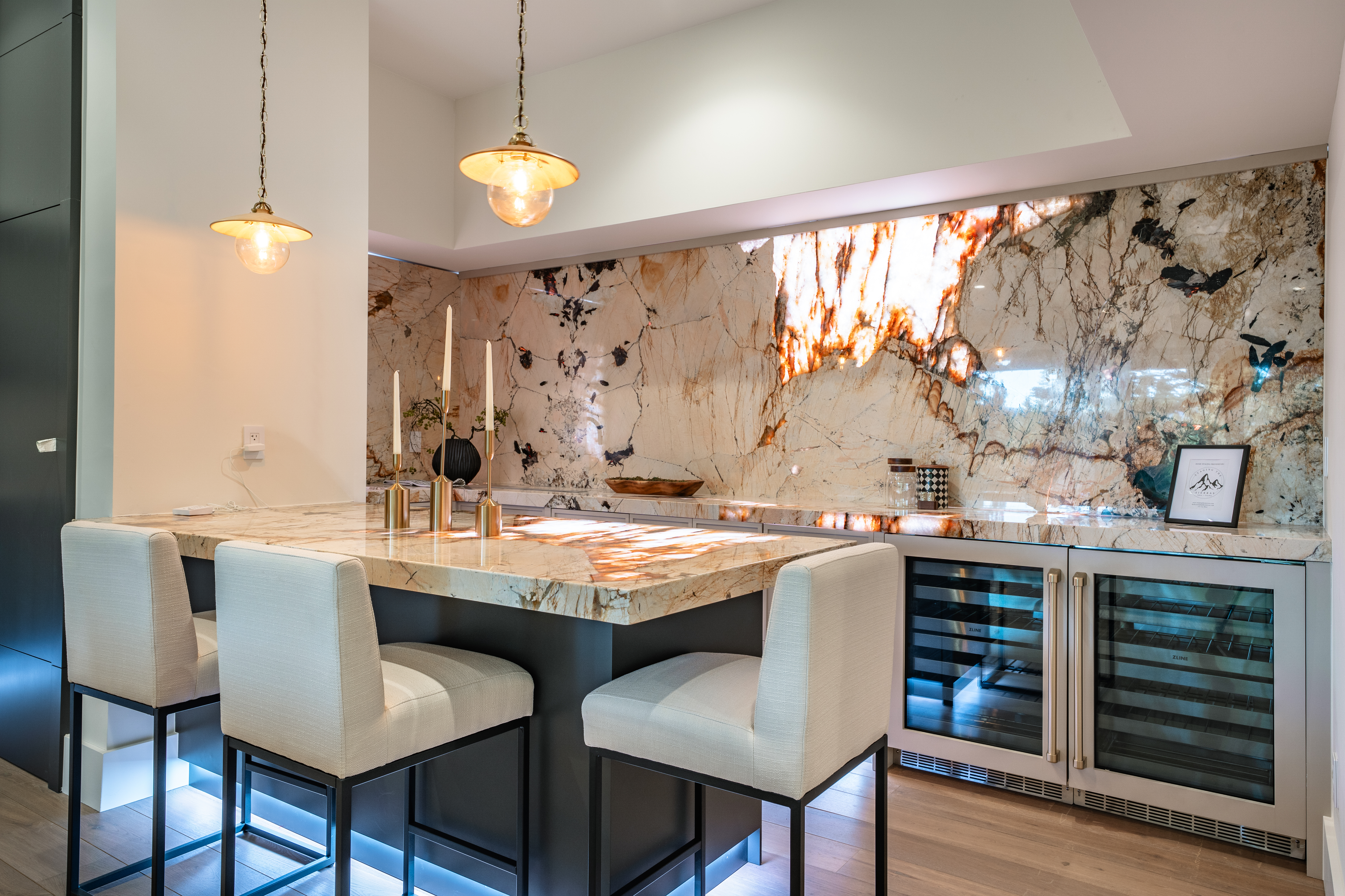 The image showcases a modern kitchen area, likely a bar or island section, featuring a stunning marble countertop and backsplash. Three contemporary bar stools with light upholstery provide seating. The design incorporates under-cabinet lighting and dual wine refrigerators, adding an upscale touch suitable for entertaining.