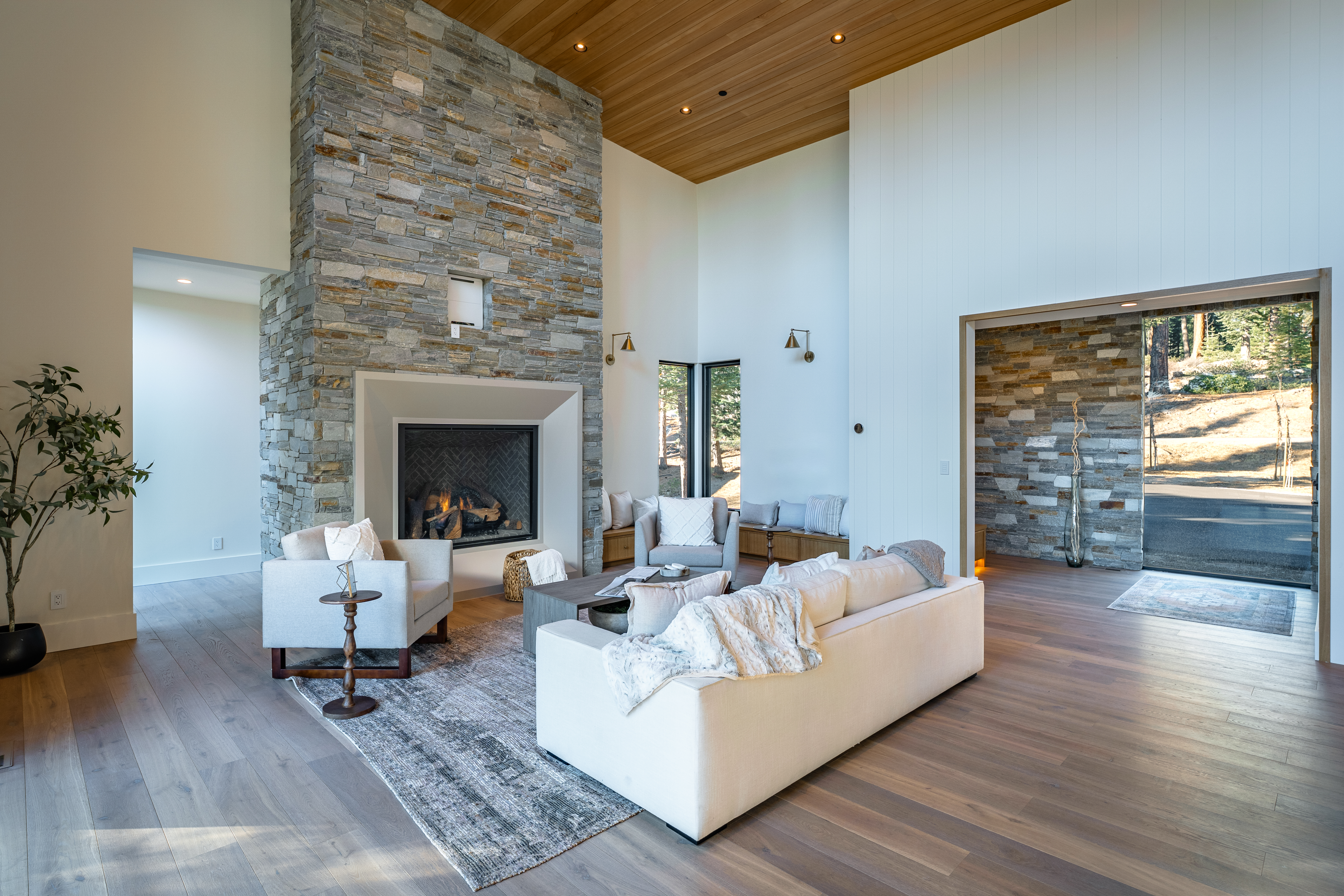 This is a bright and spacious living room featuring a stone fireplace, modern furniture, and light wood flooring. The room has high ceilings with wood paneling and ample natural light. The design is contemporary and inviting, making it an ideal space for relaxation and entertaining.