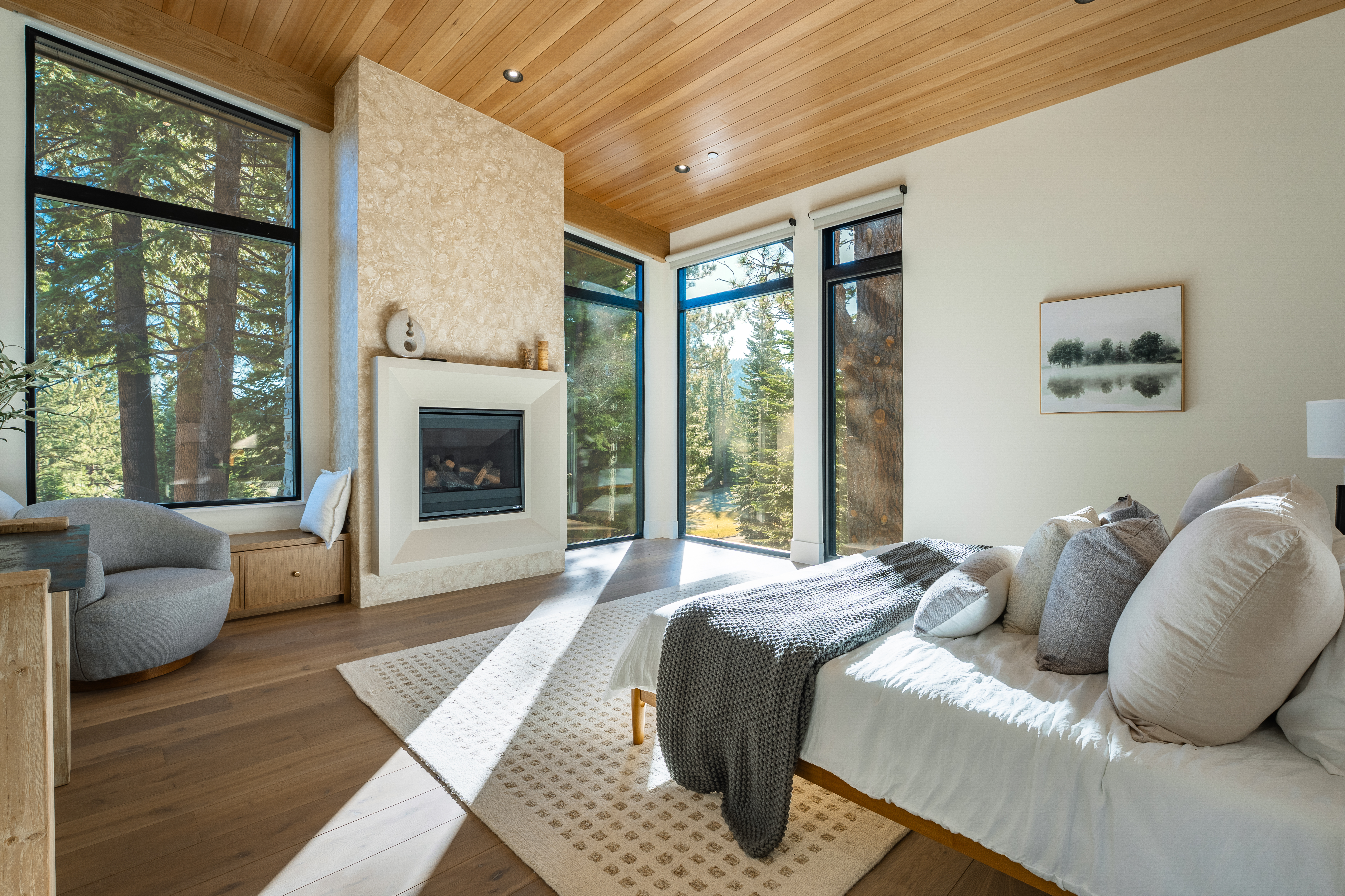 This primary bedroom boasts a modern design with natural light flooding in through large windows overlooking the forest. The room features a fireplace with a stone facade, a light-toned hardwood ceiling, and a neutral color palette, creating a serene and inviting atmosphere. A comfortable bed with plush pillows and a soft throw blanket adds to the room's appeal.