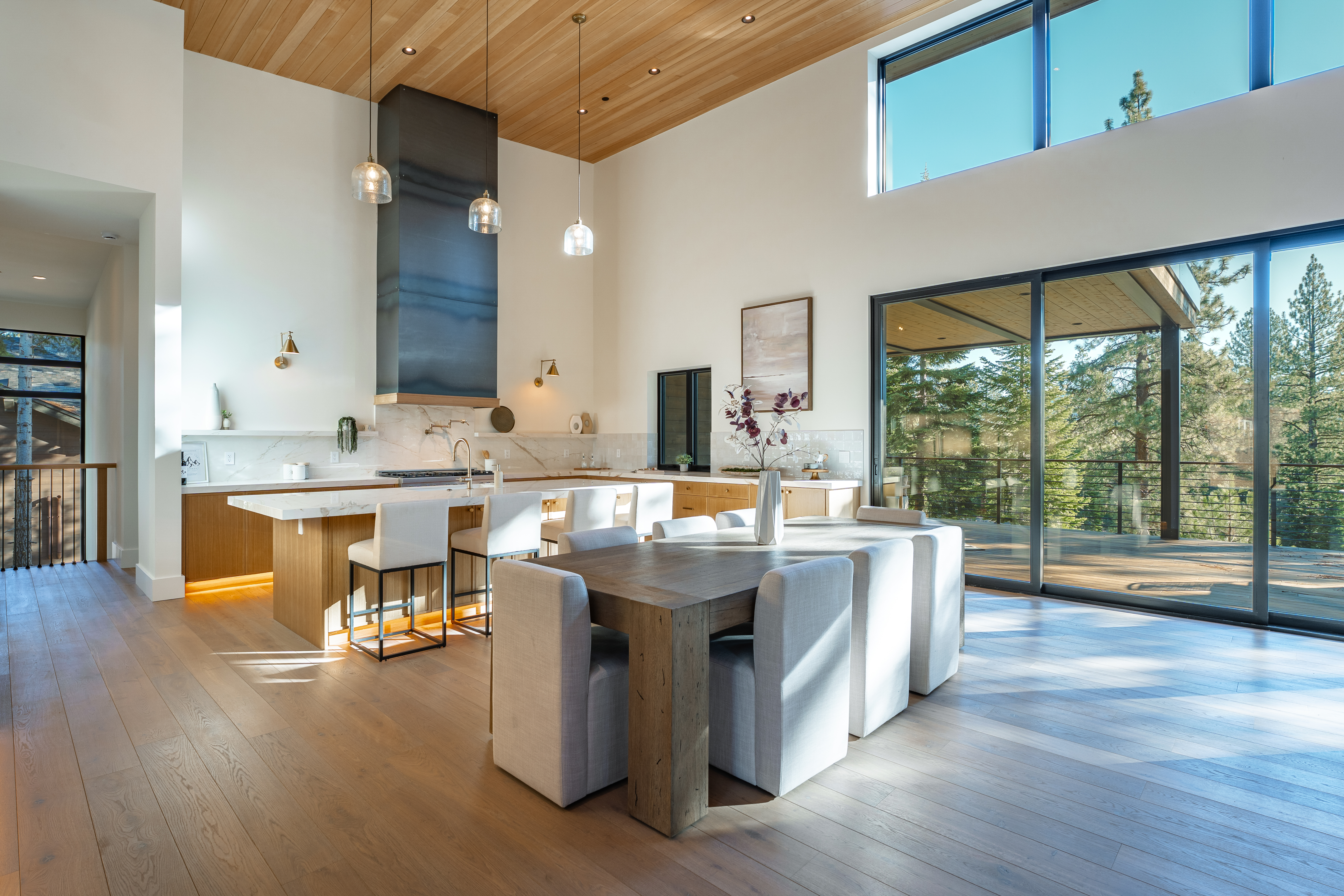 This is a bright and airy dining area situated adjacent to the kitchen, featuring modern decor and ample natural light from large sliding glass doors leading to an outdoor deck. The room includes a wooden dining table surrounded by upholstered chairs, creating a comfortable and stylish space. The high ceilings and light-toned hardwood floors add to the open and spacious feel, making it an inviting place for dining and entertaining.