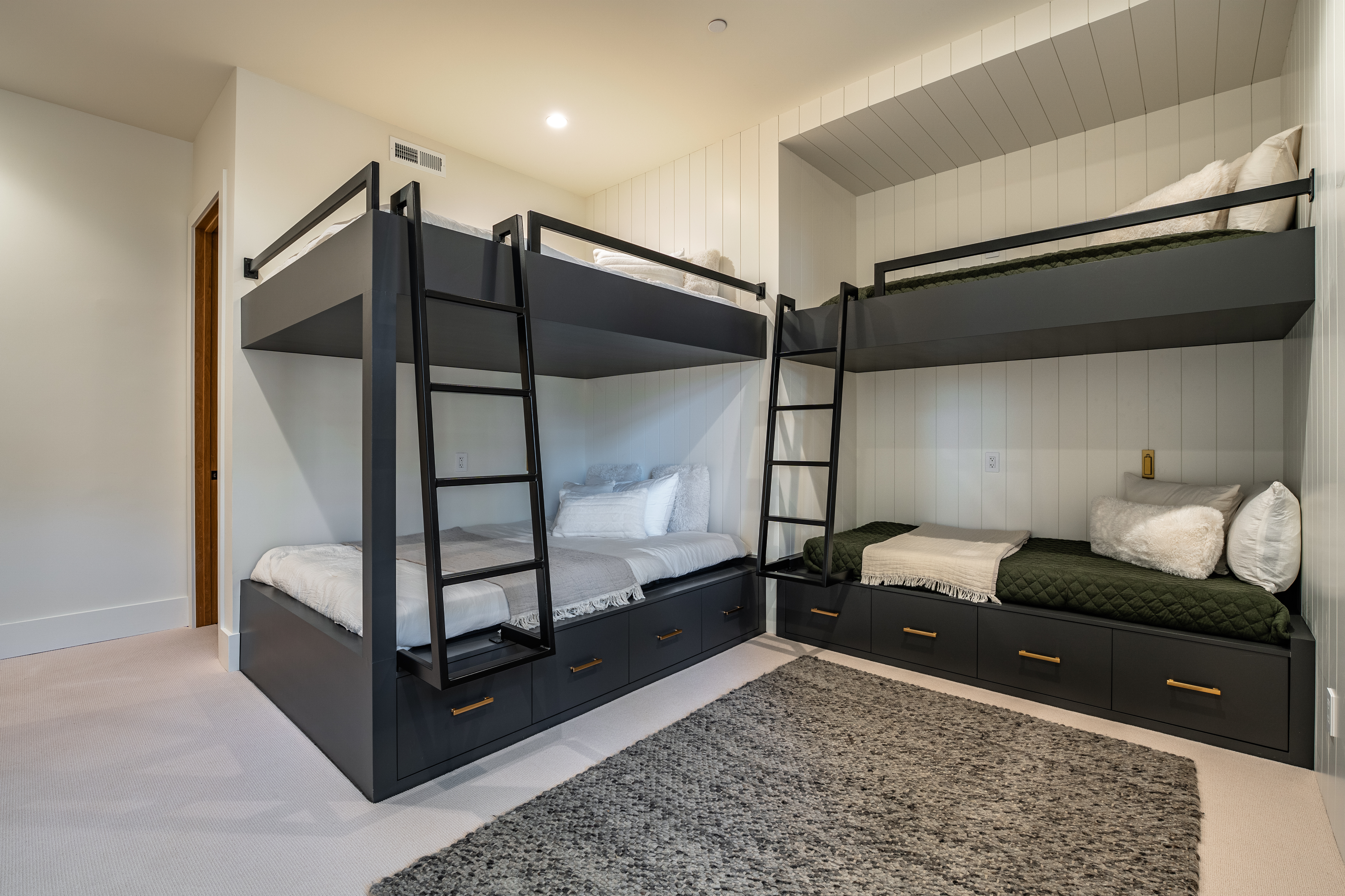 The image showcases a bedroom with two sets of modern bunk beds, each featuring a dark gray frame and drawers underneath. The beds are neatly made, and a gray patterned rug adds warmth to the carpeted floor. The room has a clean and contemporary aesthetic, perfect for a children's or guest bedroom.
