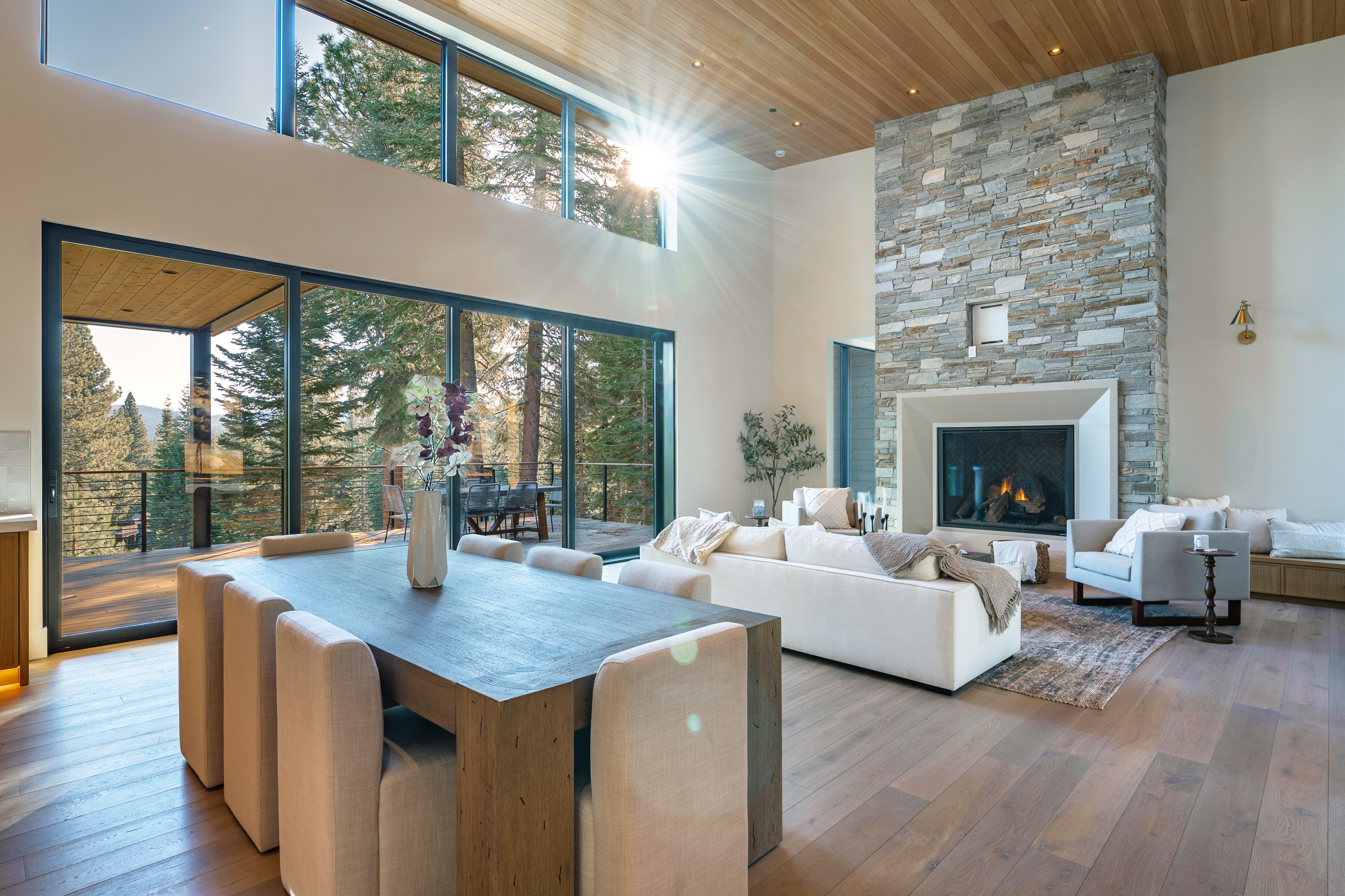 This spacious living room features a stone fireplace, hardwood floors, and large windows that bring in ample natural light and views of the surrounding landscape. The room also incorporates a dining area adding versatility to the space. Modern design elements are balanced by natural materials creating a warm and inviting atmosphere.