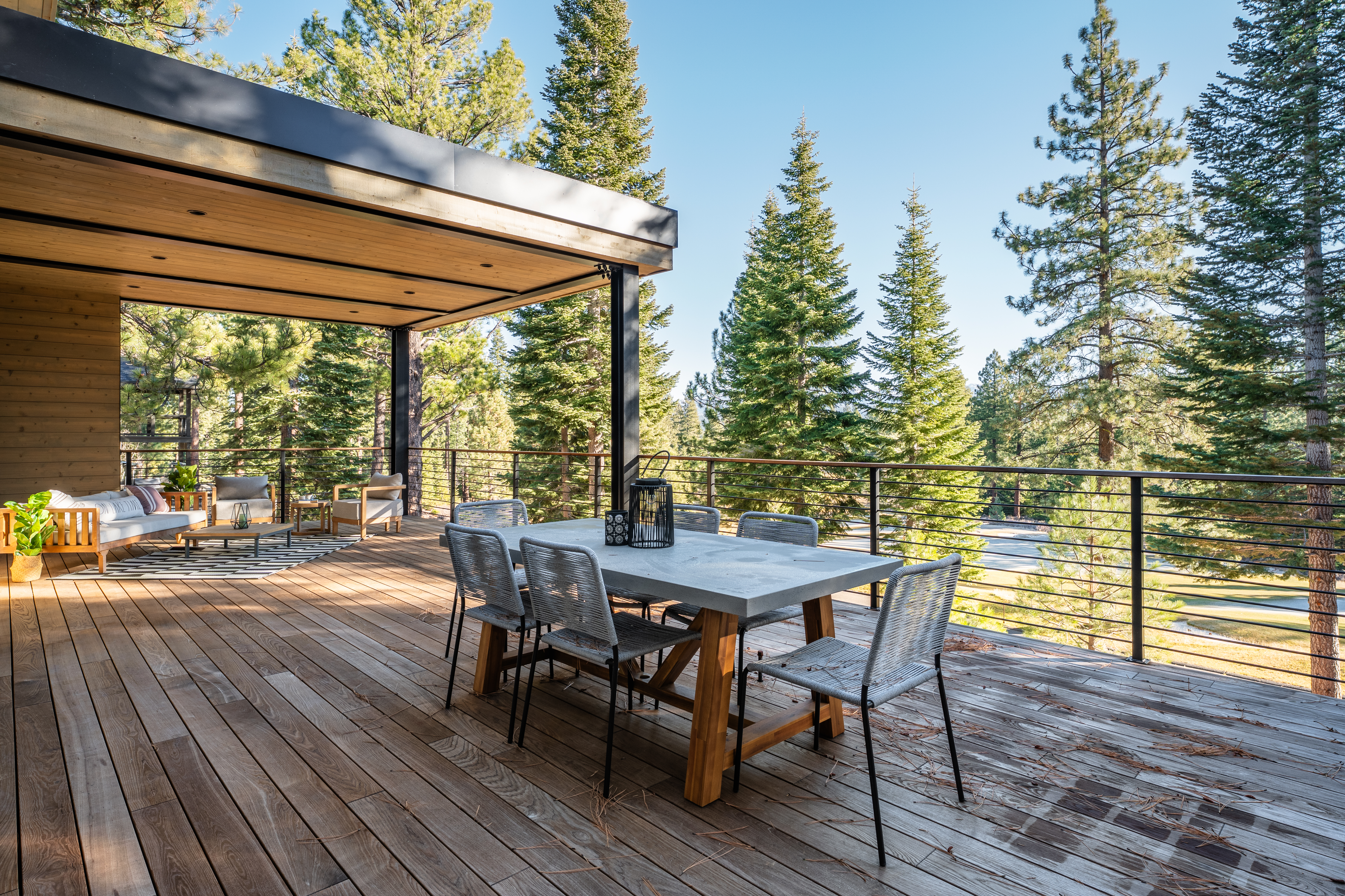 This image showcases a spacious outdoor deck area, perfect for entertaining or relaxing. The deck features a dining table with chairs and a comfortable lounge area with a sofa and chairs, creating distinct zones for different activities. The backdrop of tall trees adds to the natural and serene ambiance of the space.