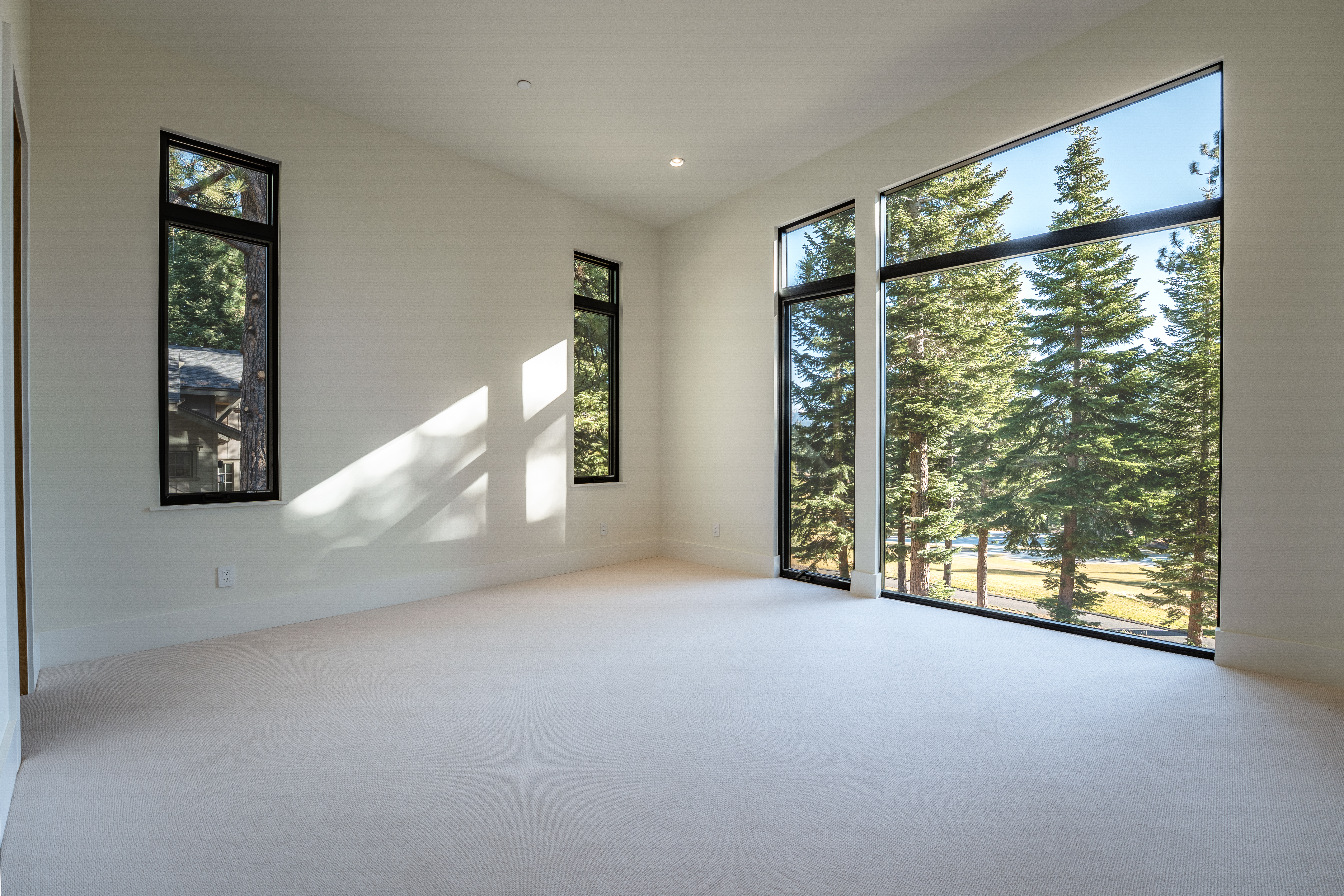 This is a bright and airy bedroom featuring large windows with views of the surrounding trees and possibly a lake. The room is carpeted in a light beige, and the walls are painted a clean white. Natural light floods the space, creating a tranquil and inviting atmosphere.