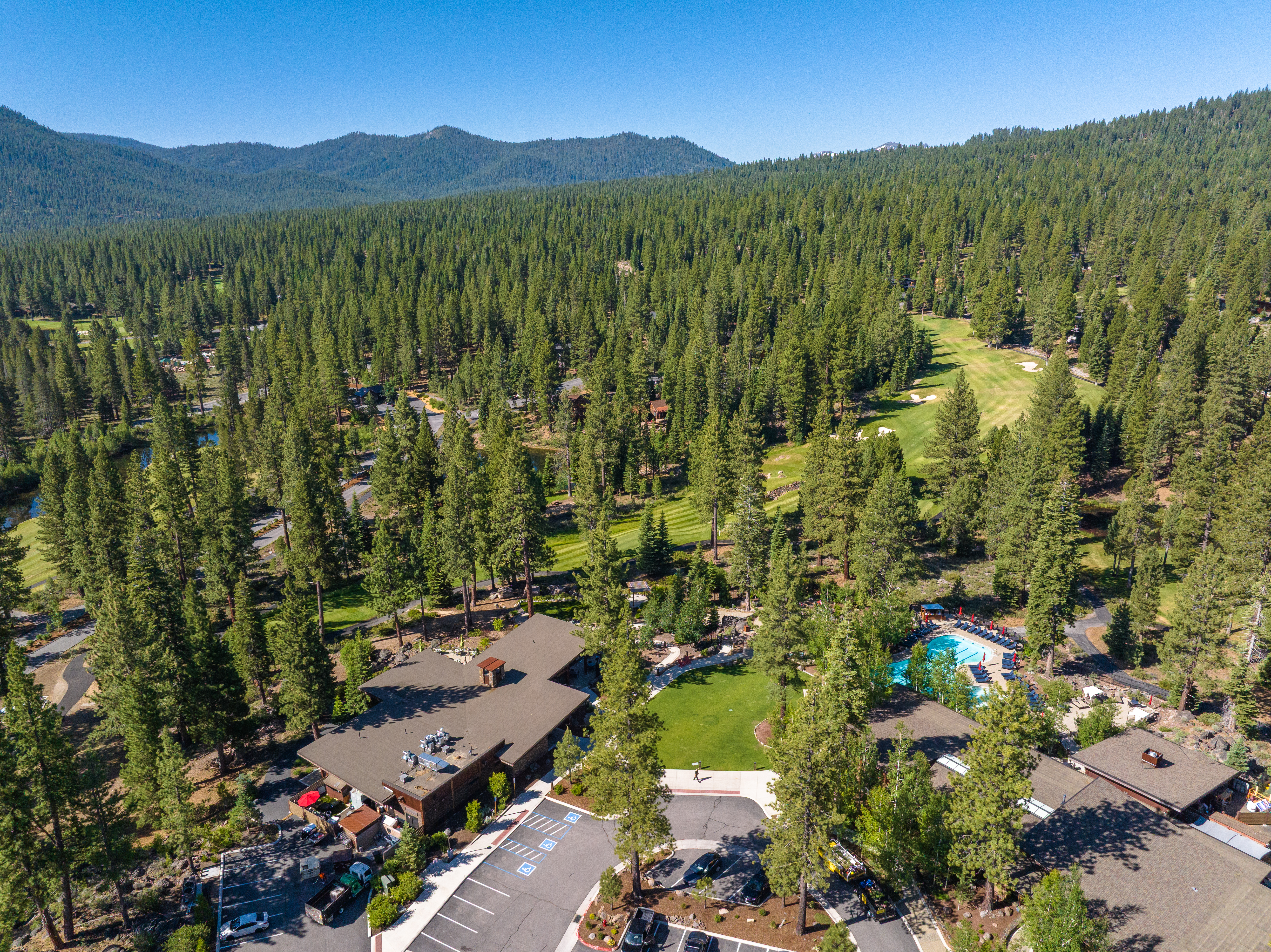 This is an aerial view showcasing a property surrounded by tall pine trees and a lush golf course. The roof of the building is visible, as well as a parking lot with parked cars. A pool area with lounge chairs offers an amenity for residents/guests.