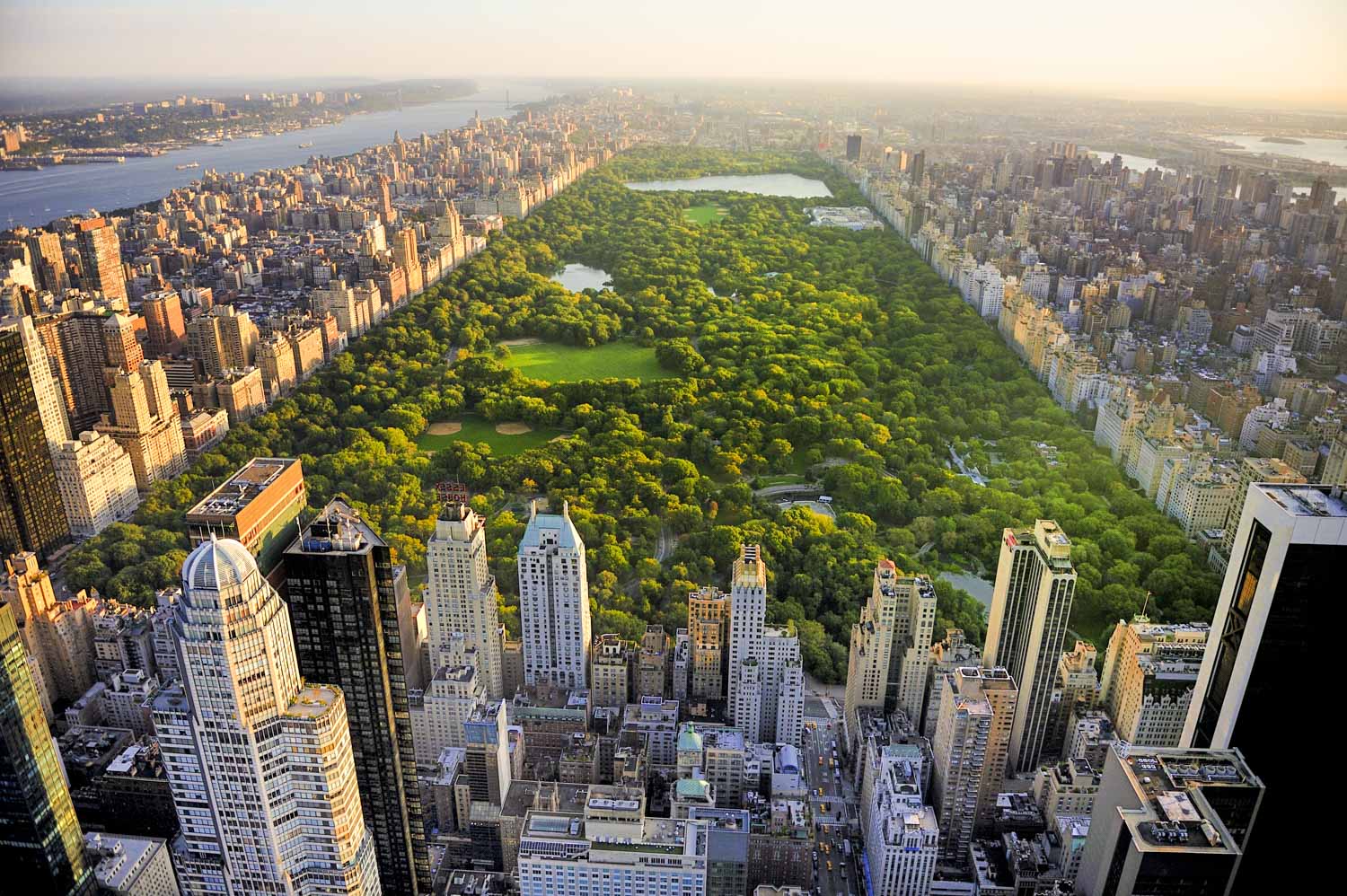 An aerial view captures the expansive landscape of Central Park in New York City, bordered by dense rows of skyscrapers. The image showcases the contrast between the natural green space and the surrounding urban environment. This perspective highlights the park's size and central location within the city.