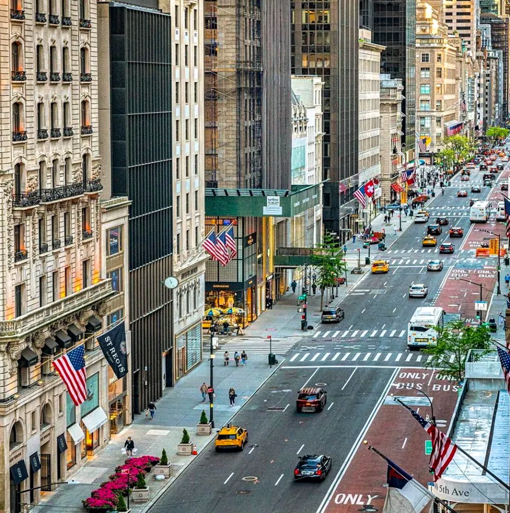 This aerial view showcases a bustling city street lined with iconic buildings decked with American flags. The street is filled with yellow taxis, buses, and other vehicles, adding to the vibrant urban scene. The architecture is a mix of classic and modern styles, suggesting a prime location in a major metropolis.