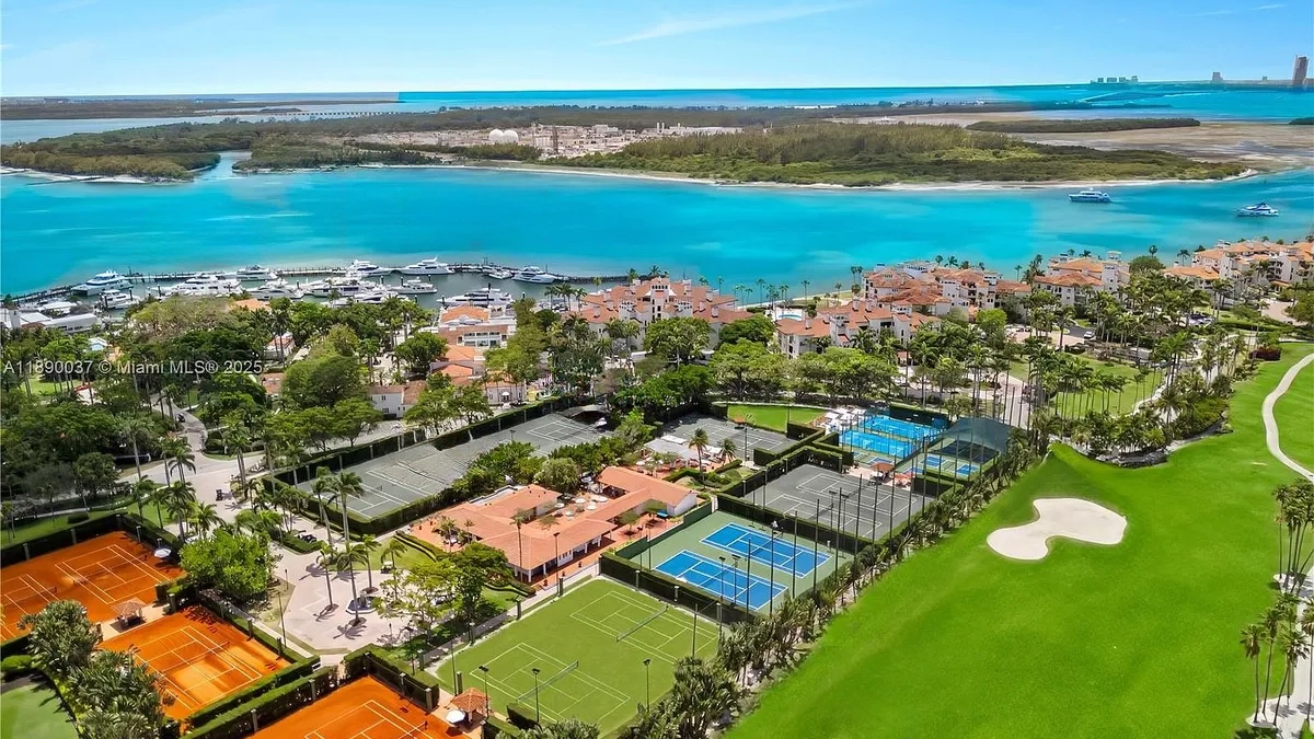 This aerial view showcases a luxurious waterfront property, featuring a well-manicured landscape with a mix of tennis courts, a golf course, and tropical trees. The neighborhood includes elegant residential buildings with red tile roofs and access to a marina filled with yachts. The turquoise water surrounds the property, creating an idyllic and exclusive setting.