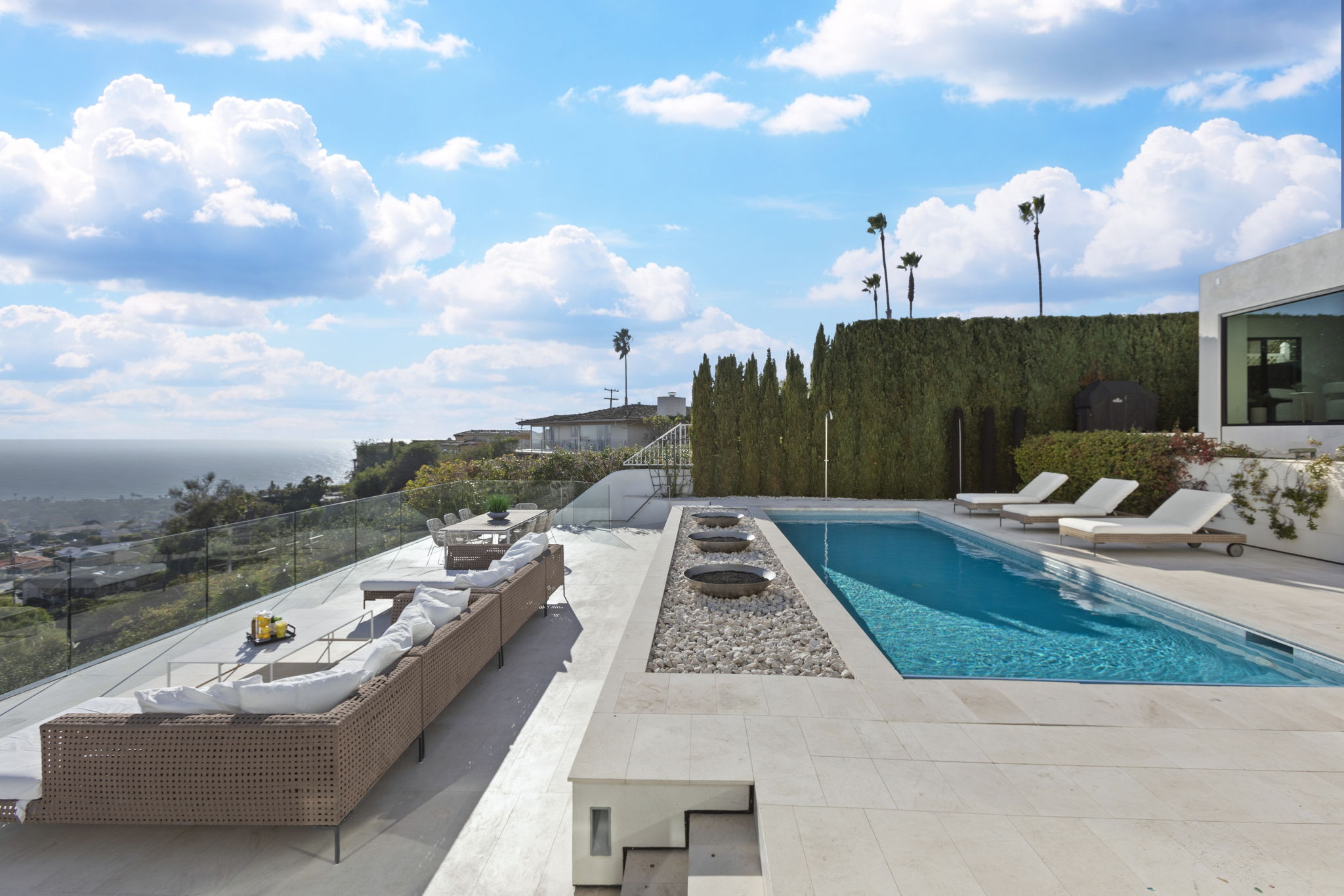 This stunning contemporary outdoor space features a rectangular swimming pool bordered by a stone-filled feature with integrated fire bowls. A spacious lounge area with modular comfortable seating and an outdoor dining area provide panoramic, breathtaking views of the coastline below. The clean, modern lines and high-end materials create an exceptionally luxurious setting perfect for entertaining or relaxation.