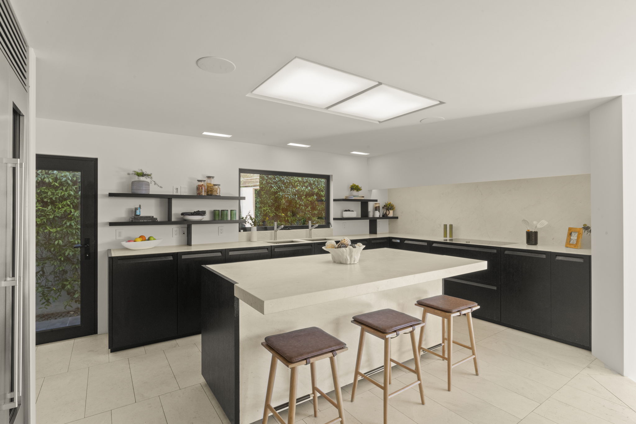 This sleek, modern kitchen features a striking high-contrast design with minimalist black cabinetry balanced by a large, light-colored stone island and matching countertops. The space is brightened by integrated ceiling lights, under-cabinet lighting, and a large window overlooking lush greenery. With its clean lines, open layout, and bar-style seating, the kitchen offers both functional workspace and an inviting atmosphere.