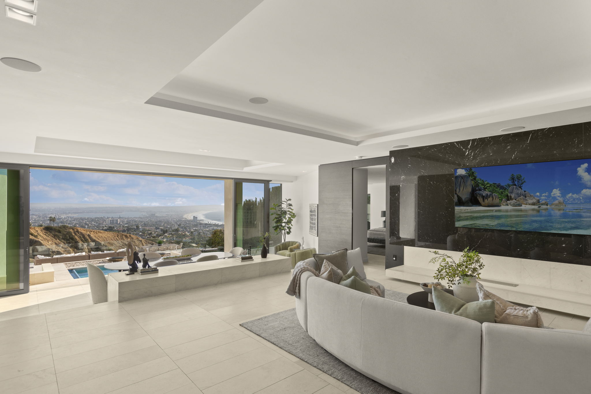 This luxurious, open-concept living room features a sweeping, curved sofa and a sleek fireplace wall accented with dark, dramatic veined stone. Large sliding glass walls completely open the space to a stunning panoramic ocean and hillside view, seamlessly blending indoor and outdoor living. The neutral color palette and clean lines create a contemporary and sophisticated atmosphere suited for high-end coastal living.