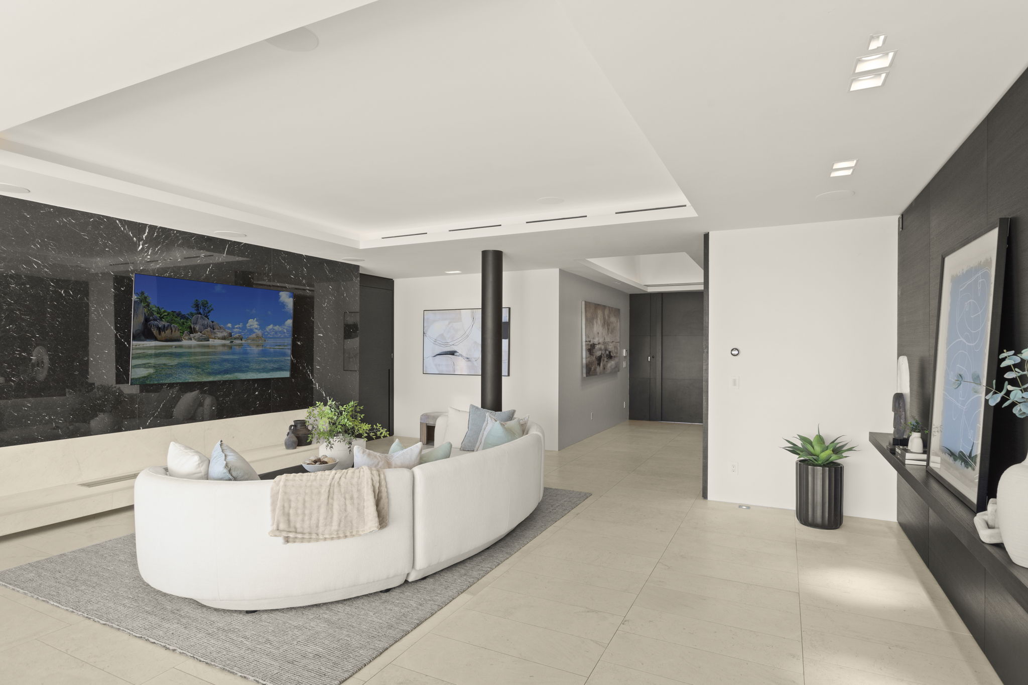 This sophisticated, contemporary living room features a sleek curved white sofa centered on a neutral area rug. A striking black marble accent wall serves as the focal point, mounting a large flat-screen television above a minimalist floating hearth. The open-concept design is accentuated by high-end finishes, recessed lighting, and elegant artwork, creating an inviting and luxurious atmosphere.