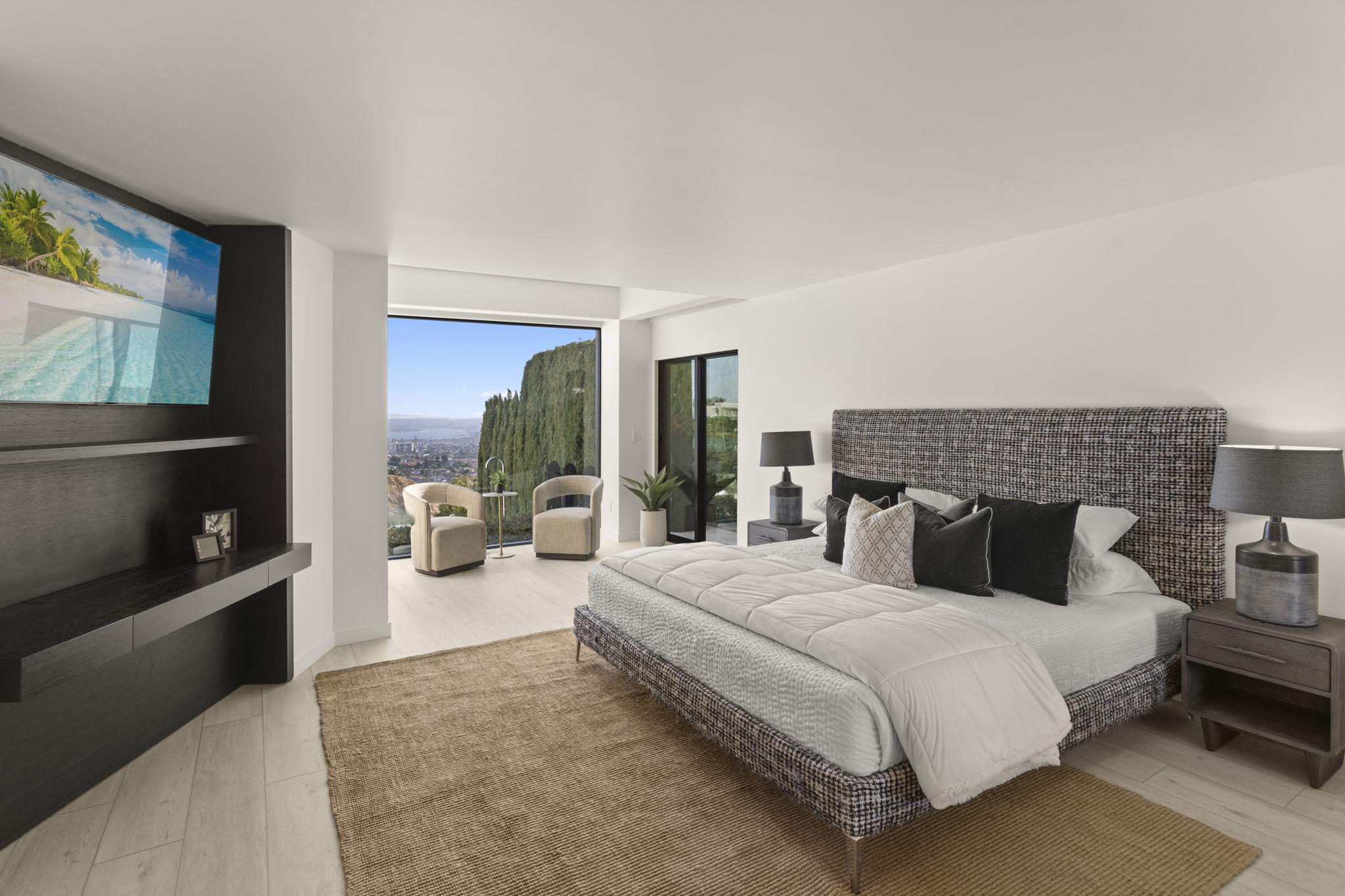 This sophisticated primary bedroom features a striking grey tweed-textured headboard and a plush, neutral-toned bedding set. The room is anchored by a large, natural fiber area rug and flows seamlessly into a private sitting area with floor-to-ceiling windows that provide panoramic views. A dark wood entertainment console and matching nightstands add a modern, grounded aesthetic to this bright and airy space.